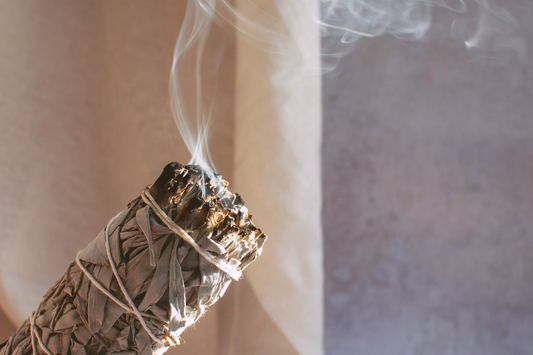 A large bundle of white sage leaves, tied into a stick with white string. The bundle has been lit and blown out and is wafting fragrant smoke across a neutral background.