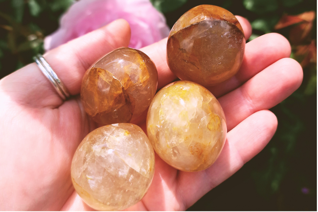 Four yellow, brown and clear coloured translucent golden healer quartz polished rounded tumblestones sit in the palm of a hand. A pink rose grows in the background.