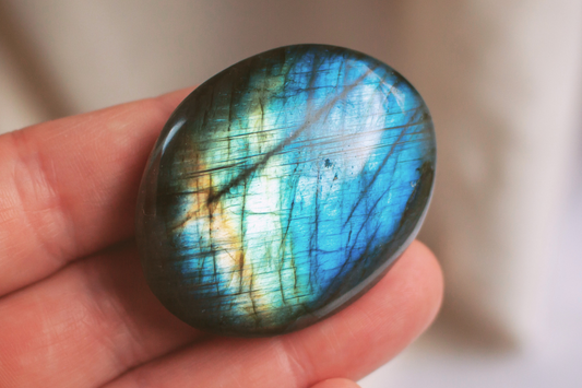 A hand holding a bright polished oval shaped labradorite crystal palmstone, displaying flashes of blue, green and gold colours.