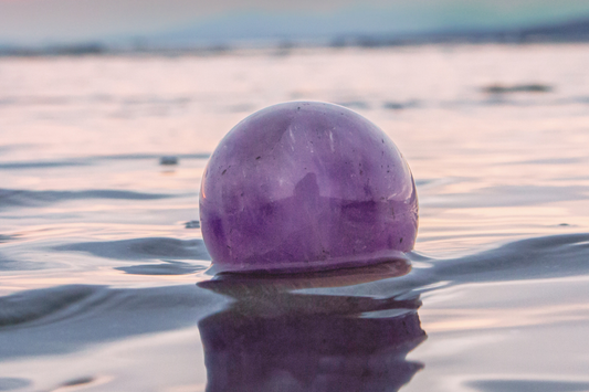 A large translucent purple amethyst sphere sits partially submerged in the ocean on a beach at sunset.