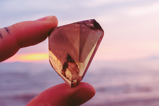 A polished and shaped natural yellow citrine crystal is being held up to the light by a finger and thumb. An ocean sunset is in the background, giving everything a pinkish glow.