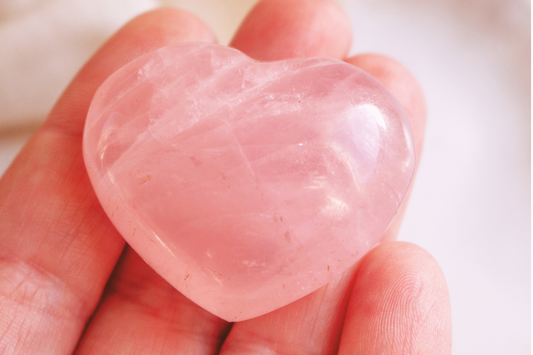 A large translucent dark pink rose quartz heart sits on the fingers of a woman's palm over a neutral beige background.