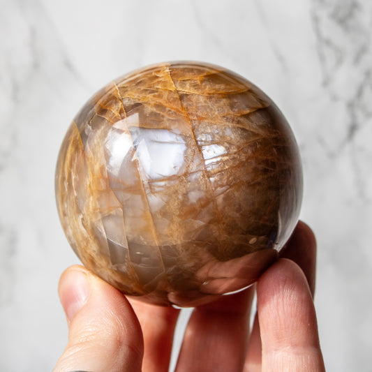 a brown crystal sphere with a bright silver flash being held in fingertips with a blurred marble white background