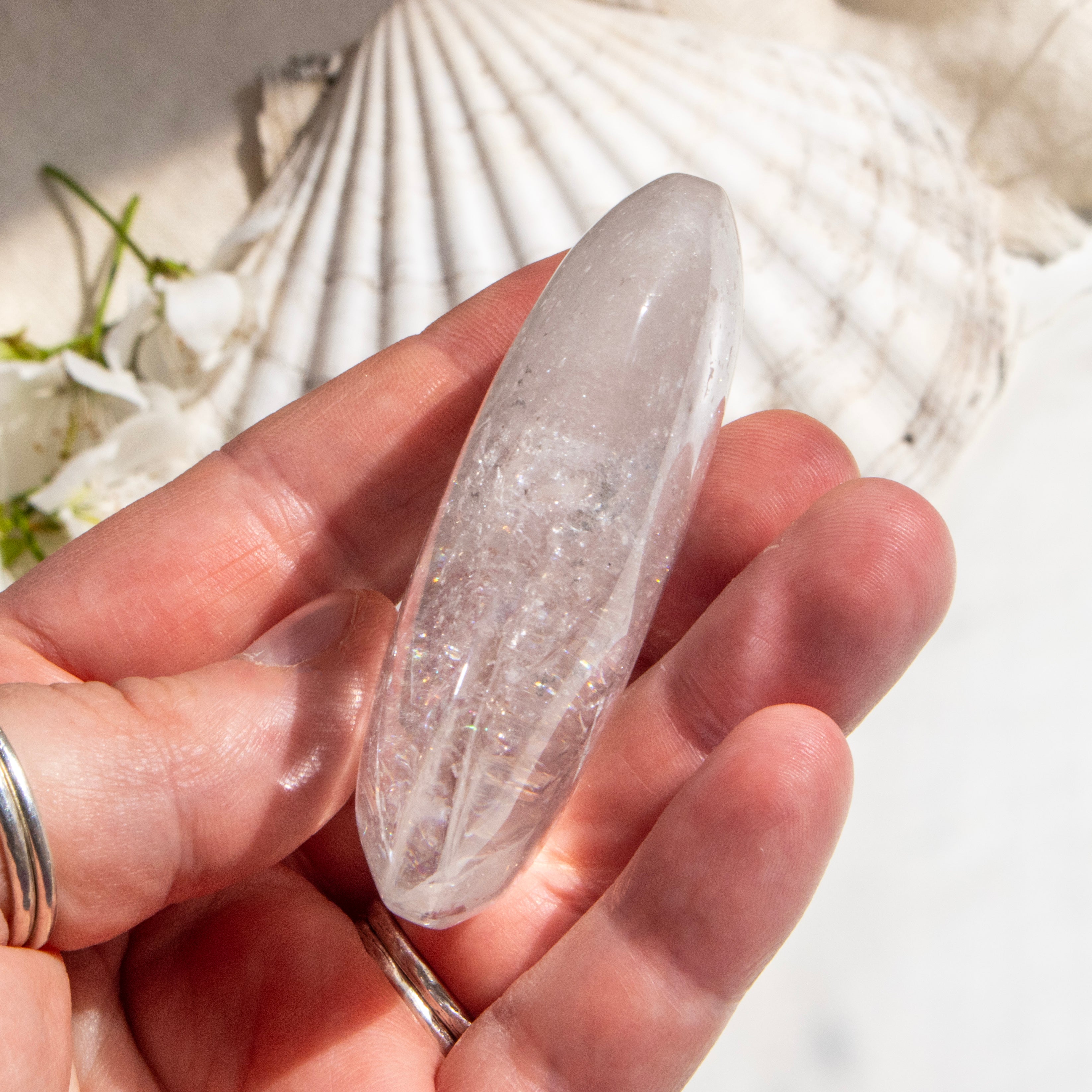 Clear crystal stone held in a hand with a shell and flowers in the background