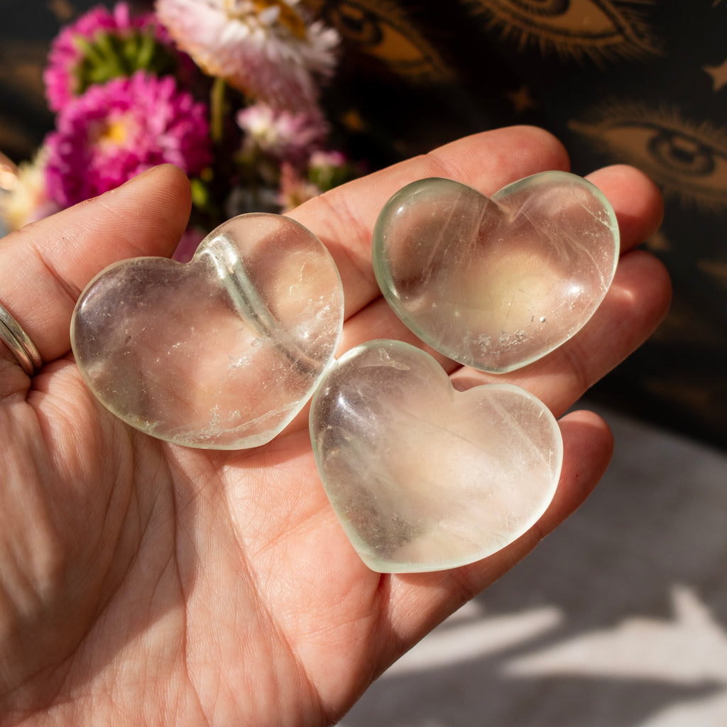 Three clear heart-shaped stones held in a hand with a blurred floral background