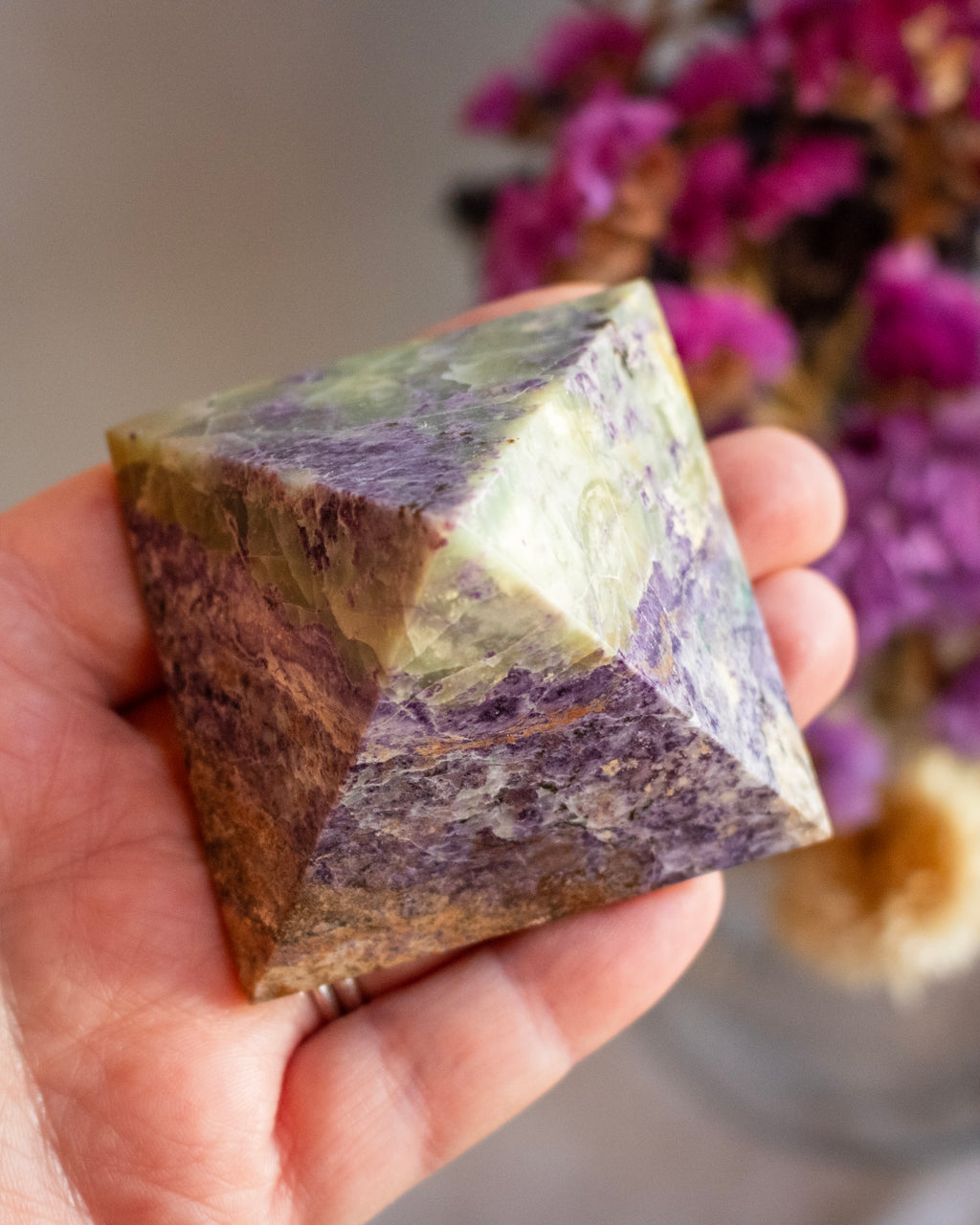 Hand holding a pyramid-shaped crystal with a blurred background of flowers
