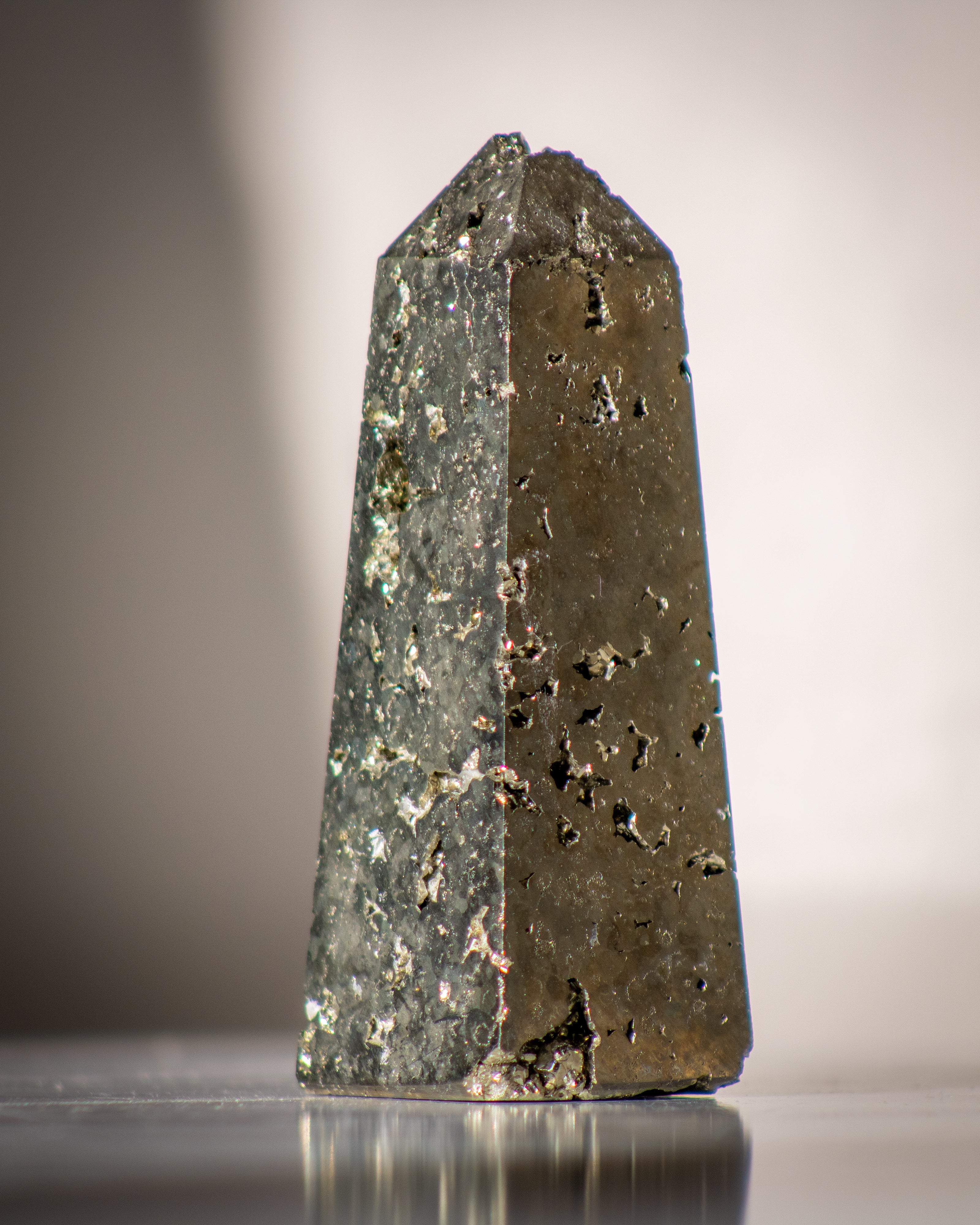 Tall, dark gold mineral crystal on a reflective surface with a neutral background