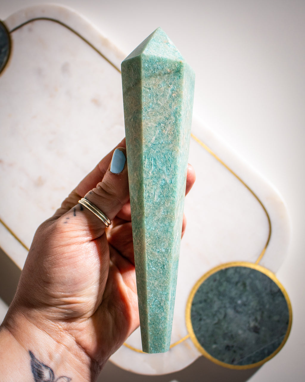 a large green amazonite crystal shaped like a long tapered wand with points at both ends being held by a hand over a white background.