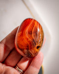 Hand holding a polished red and brown stone with a wavy pattern against a neutral background
