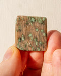 Square stone with green speckles held between fingers against a beige background