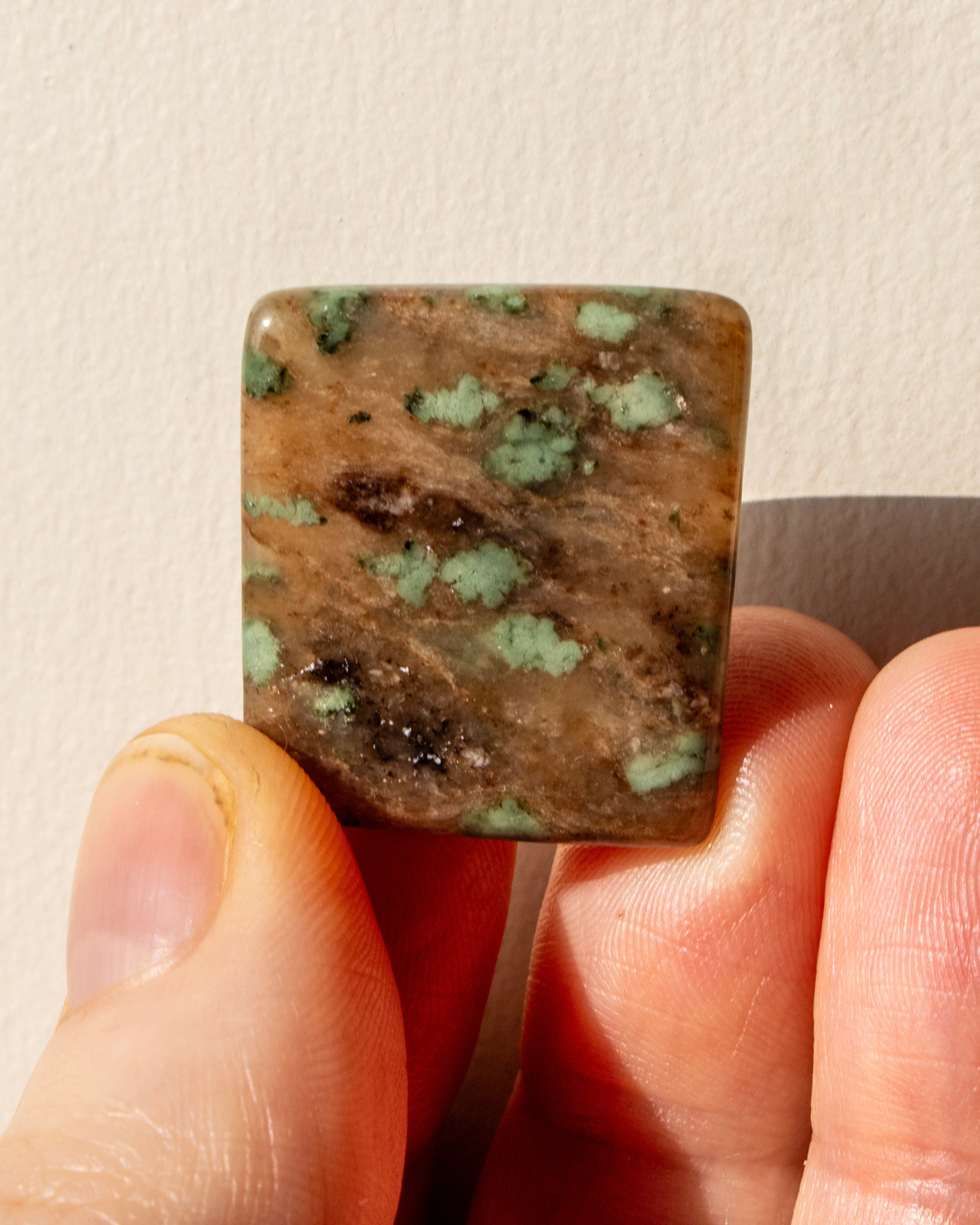 Square stone with green and brown patterns held between fingers against a neutral background
