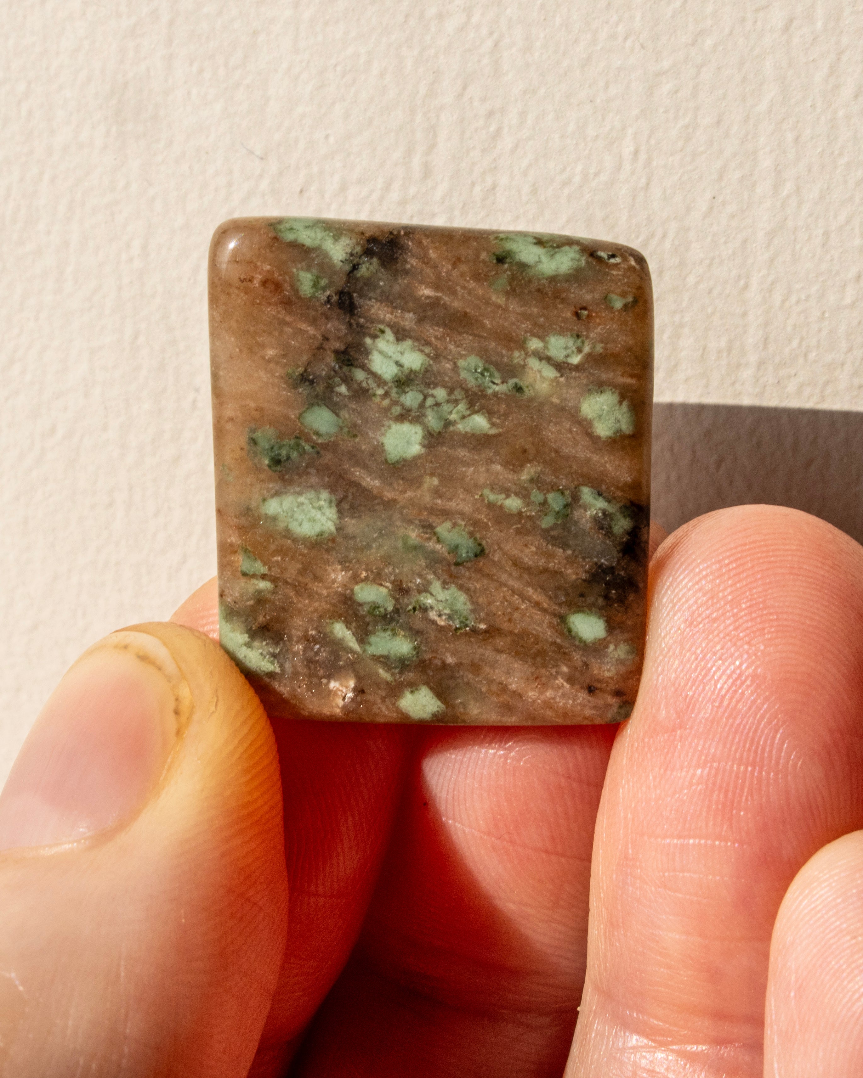 Square-shaped stone with green and brown patterns held between fingers against a neutral background