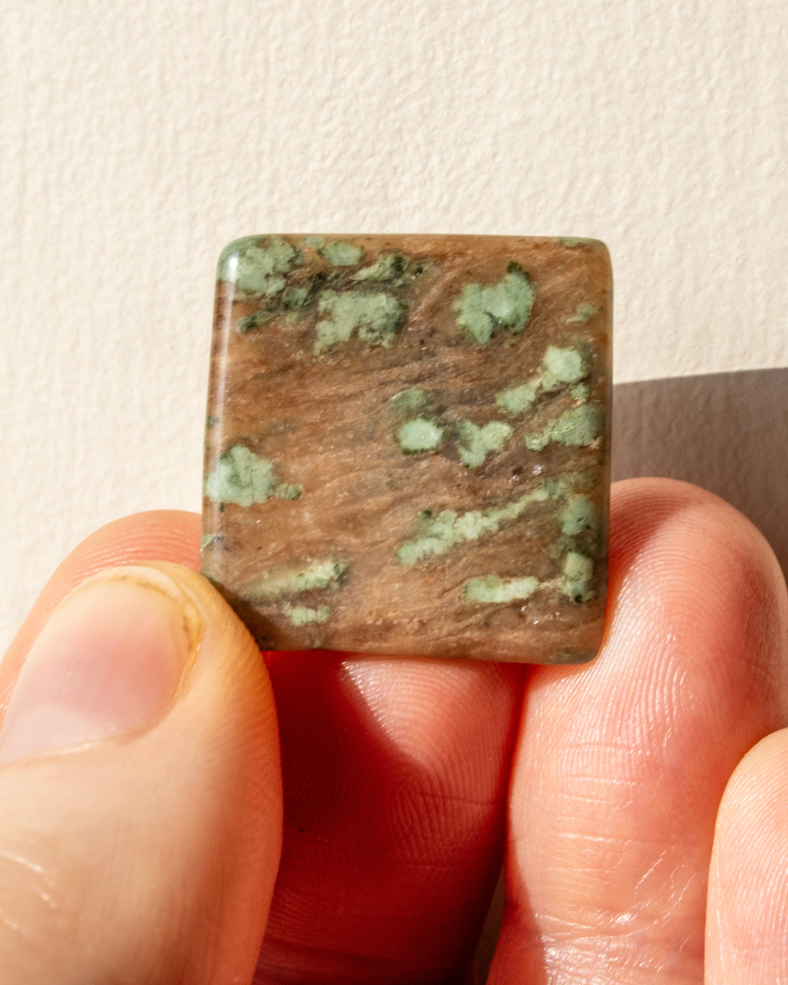 Square stone with brown and green pattern held between fingers against a neutral background
