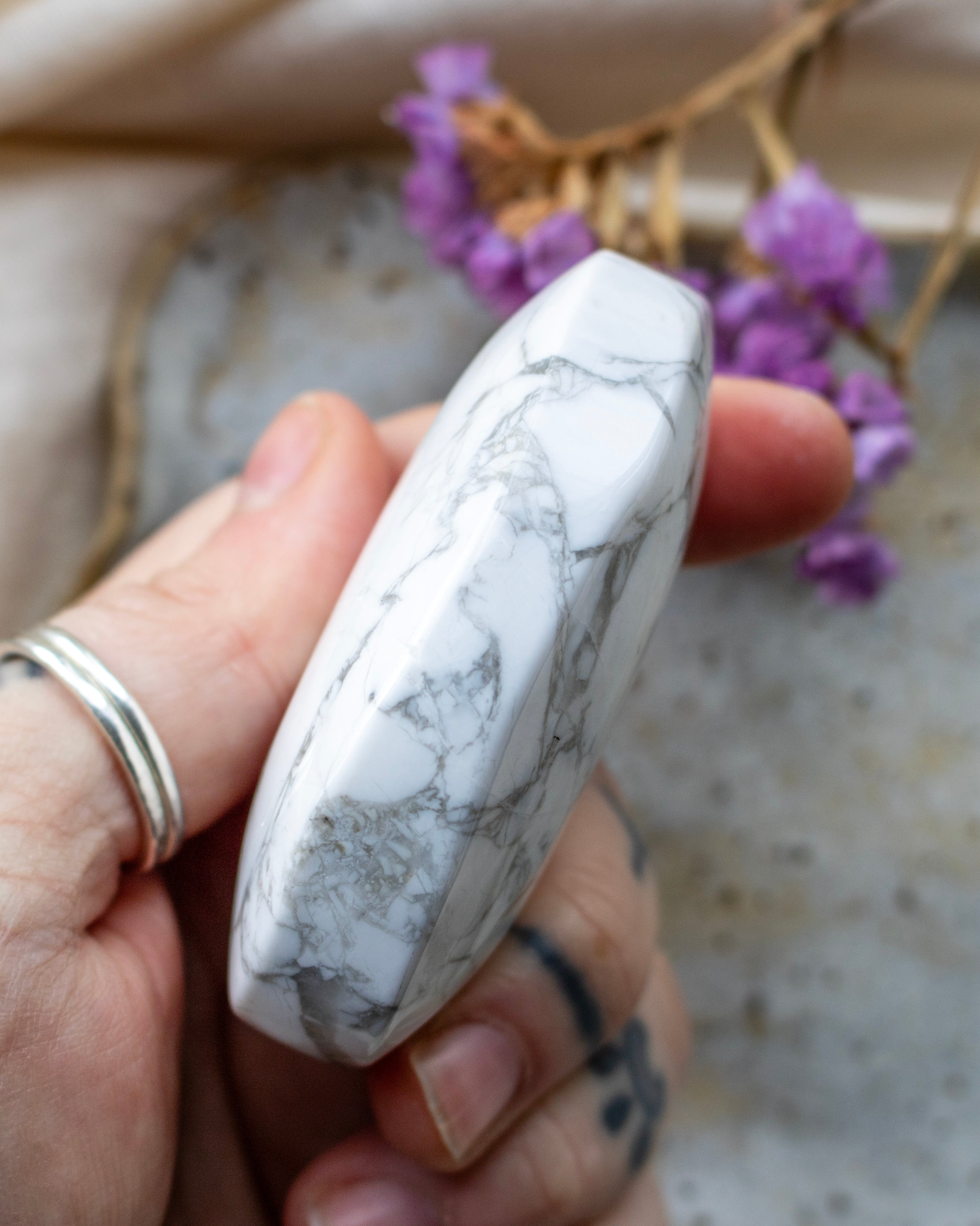 Hand holding a marbled stone with a blurred background featuring purple flowers and a beige surface.