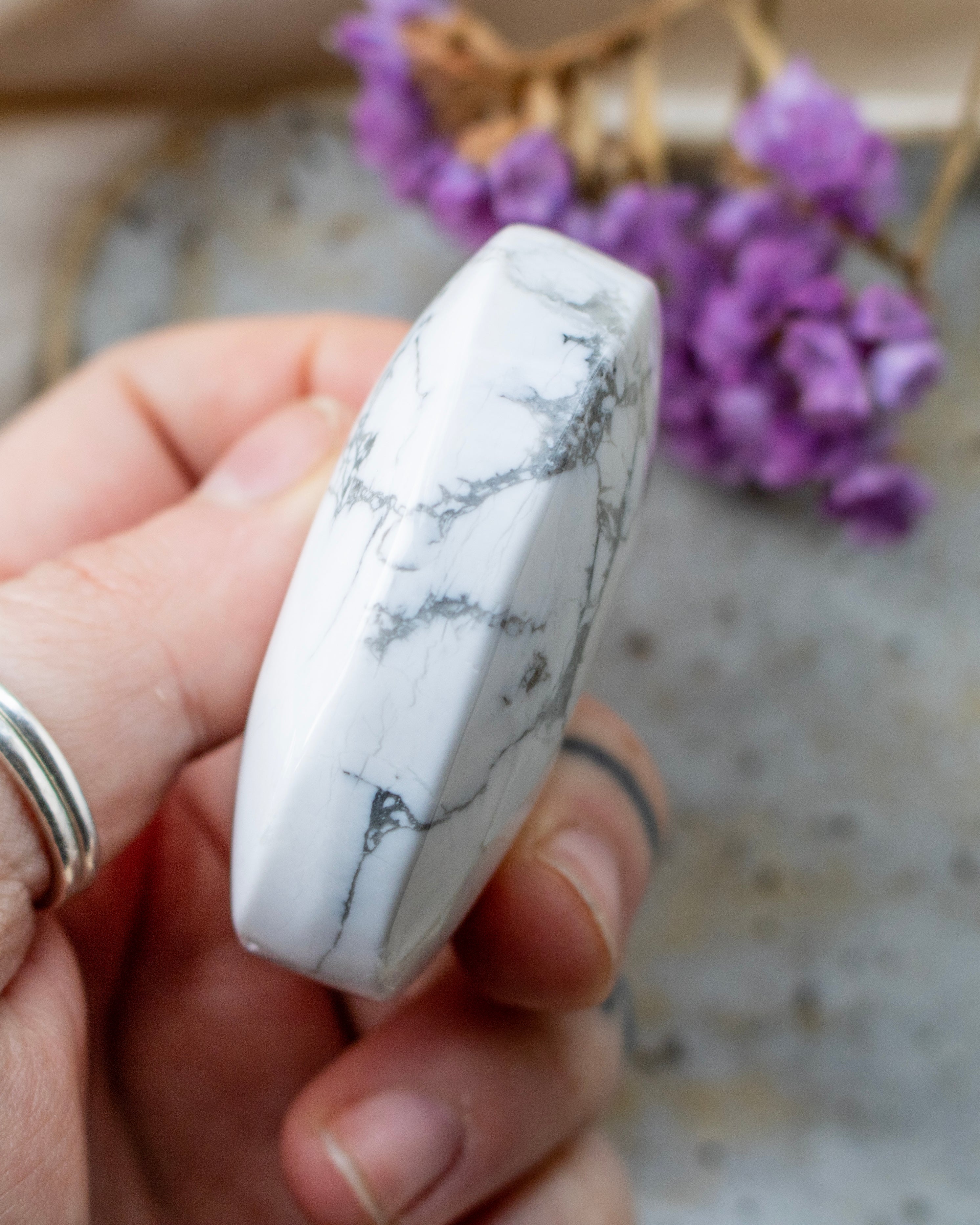 Hand holding a white stone with gray marbling, blurred purple flowers in the background