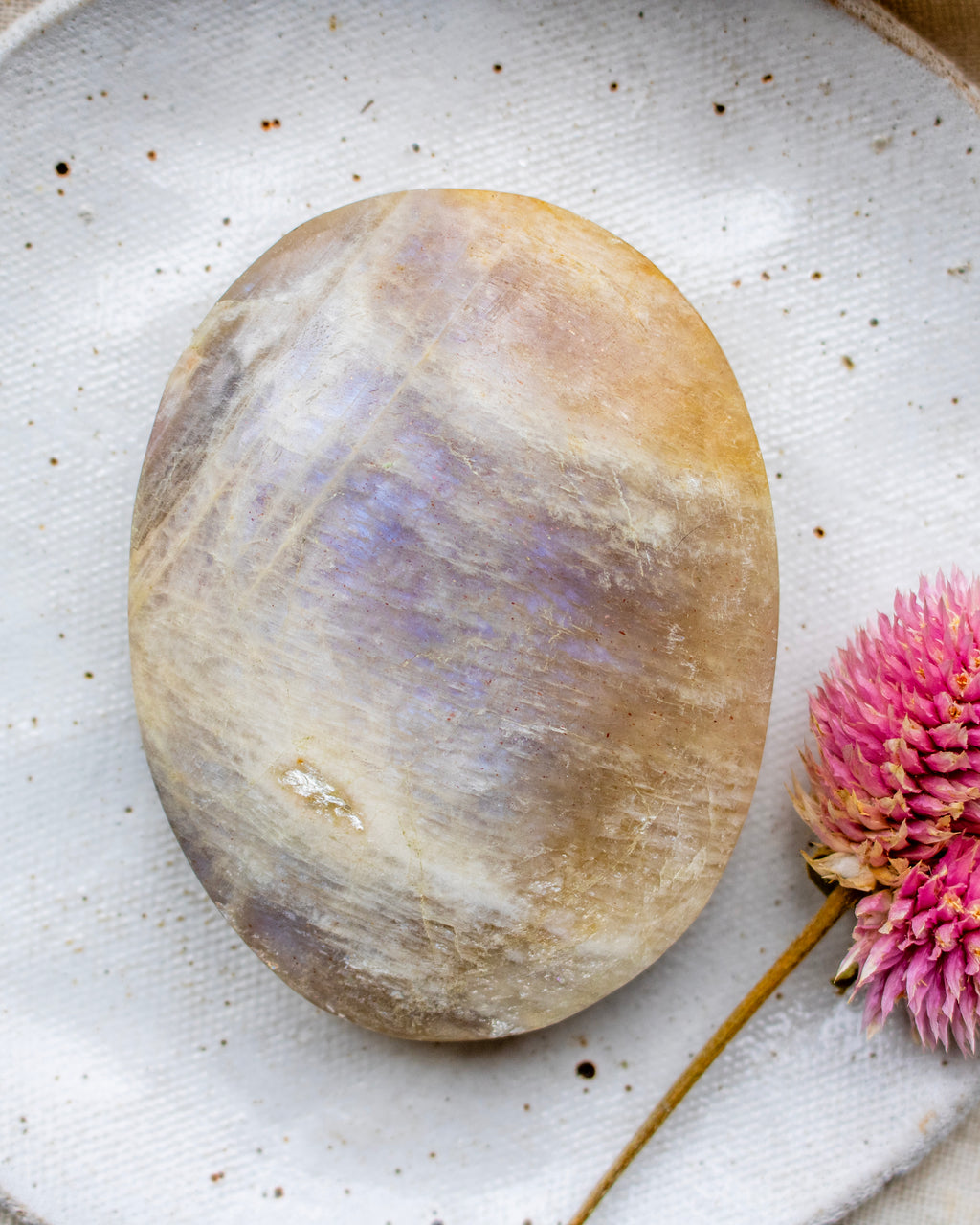 Polished stone with a rainbow-like pattern on a textured white surface with a pink flower.