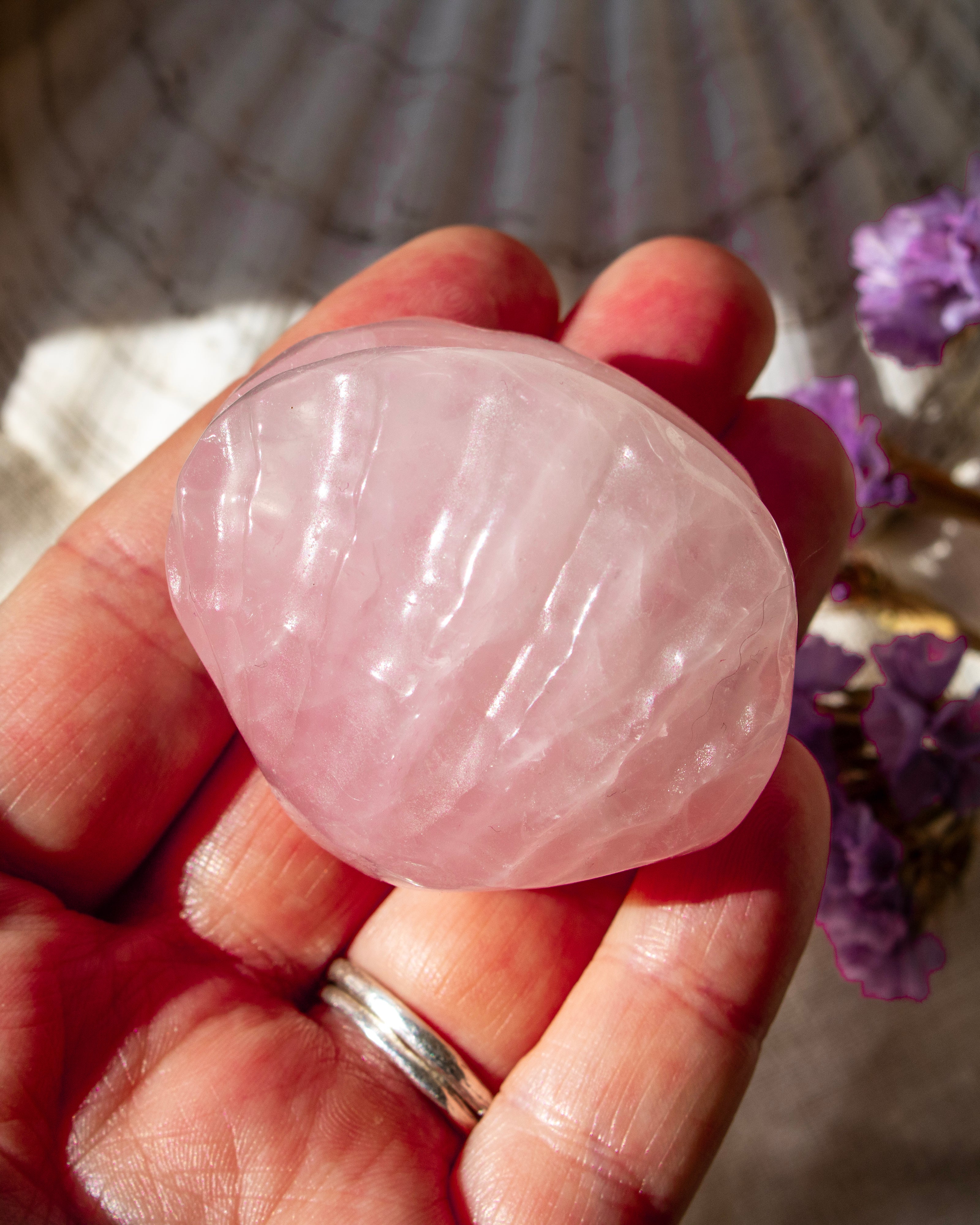 Hand holding a pink rose quartz crystal with blurred purple flowers in the background