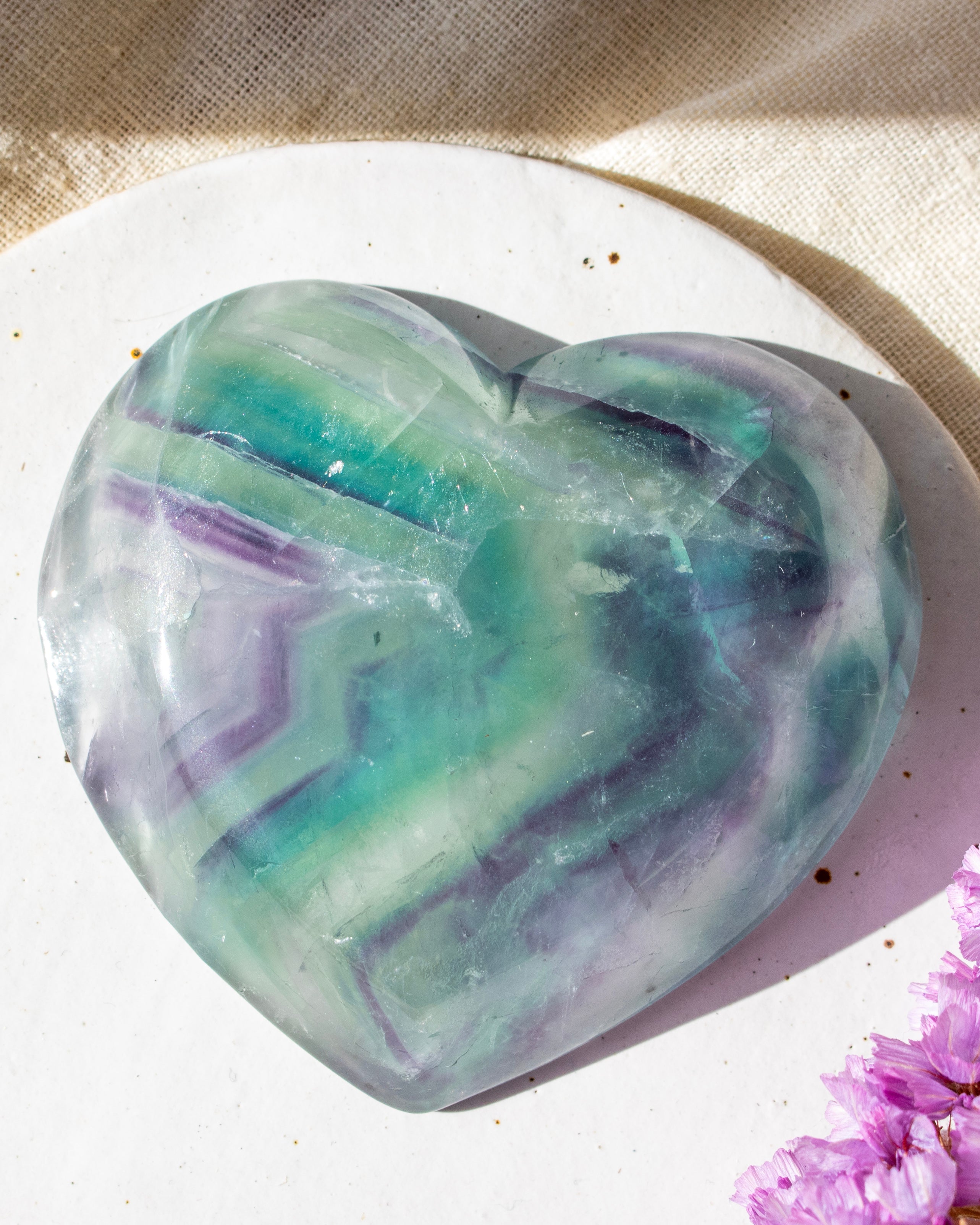 Heart-shaped green and purple crystal on a white surface with a pink flower in the corner
