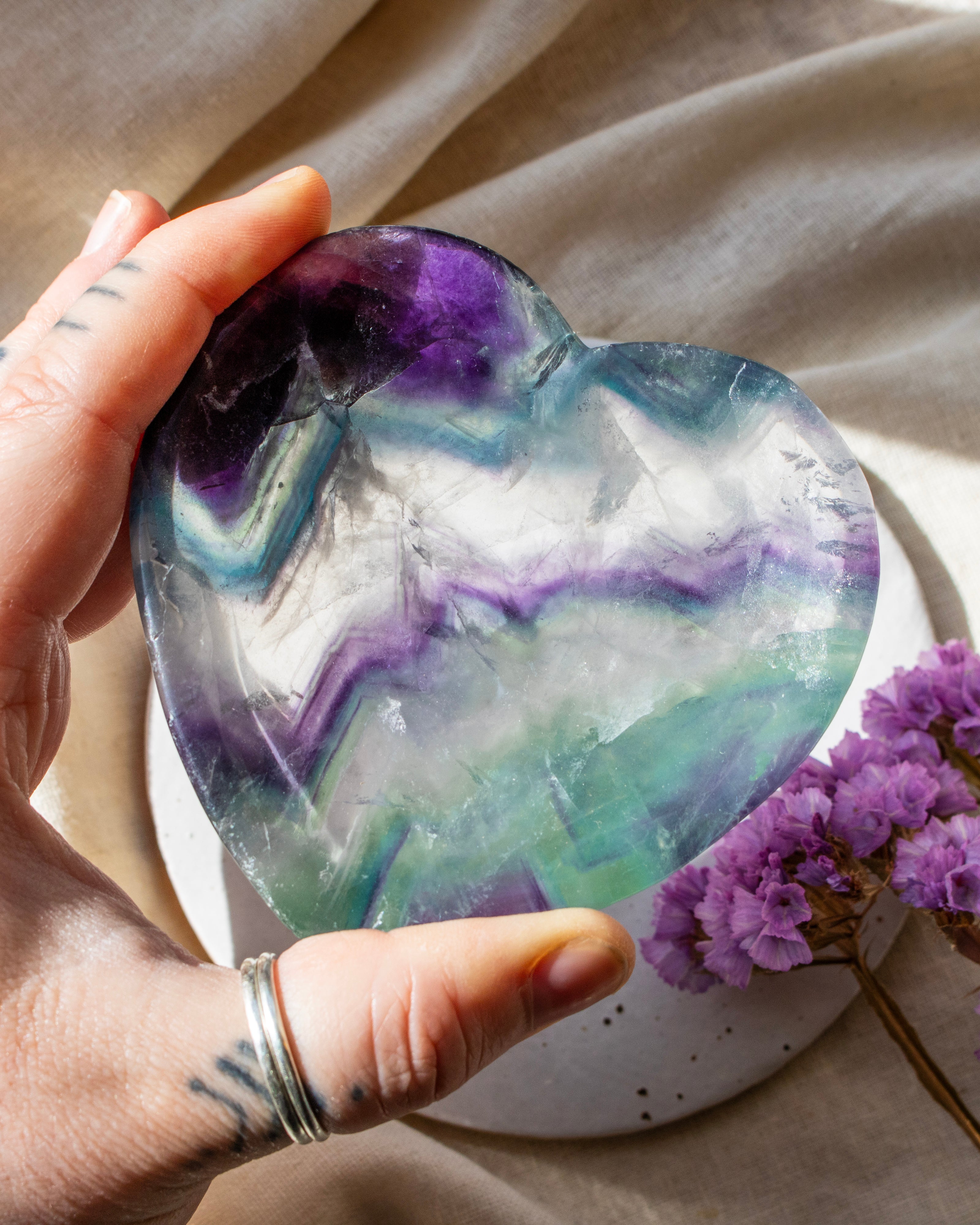 Heart-shaped crystal with purple and green colors held by a hand, with a blurred background