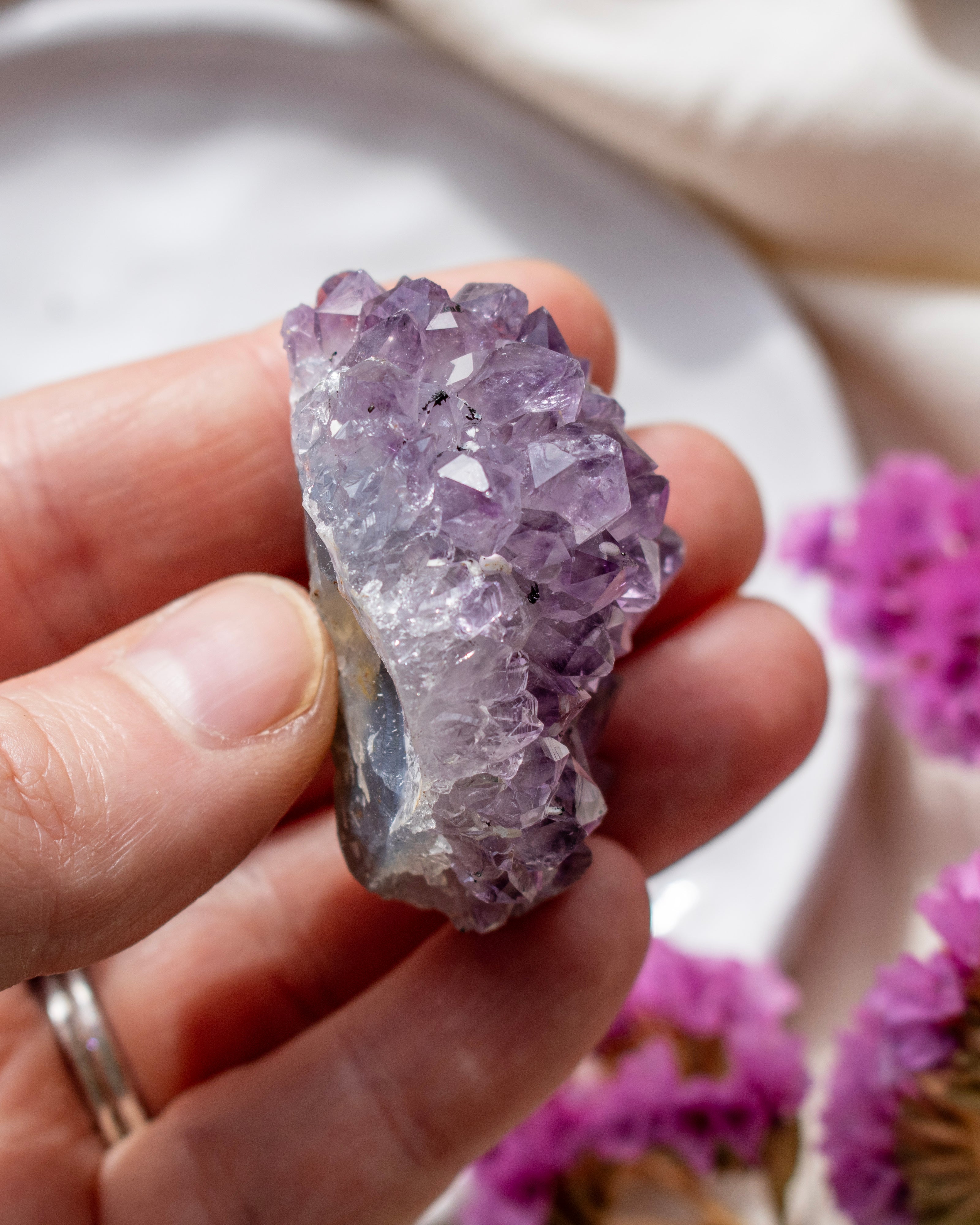 Hand holding a purple crystal with blurred pink flowers in the background