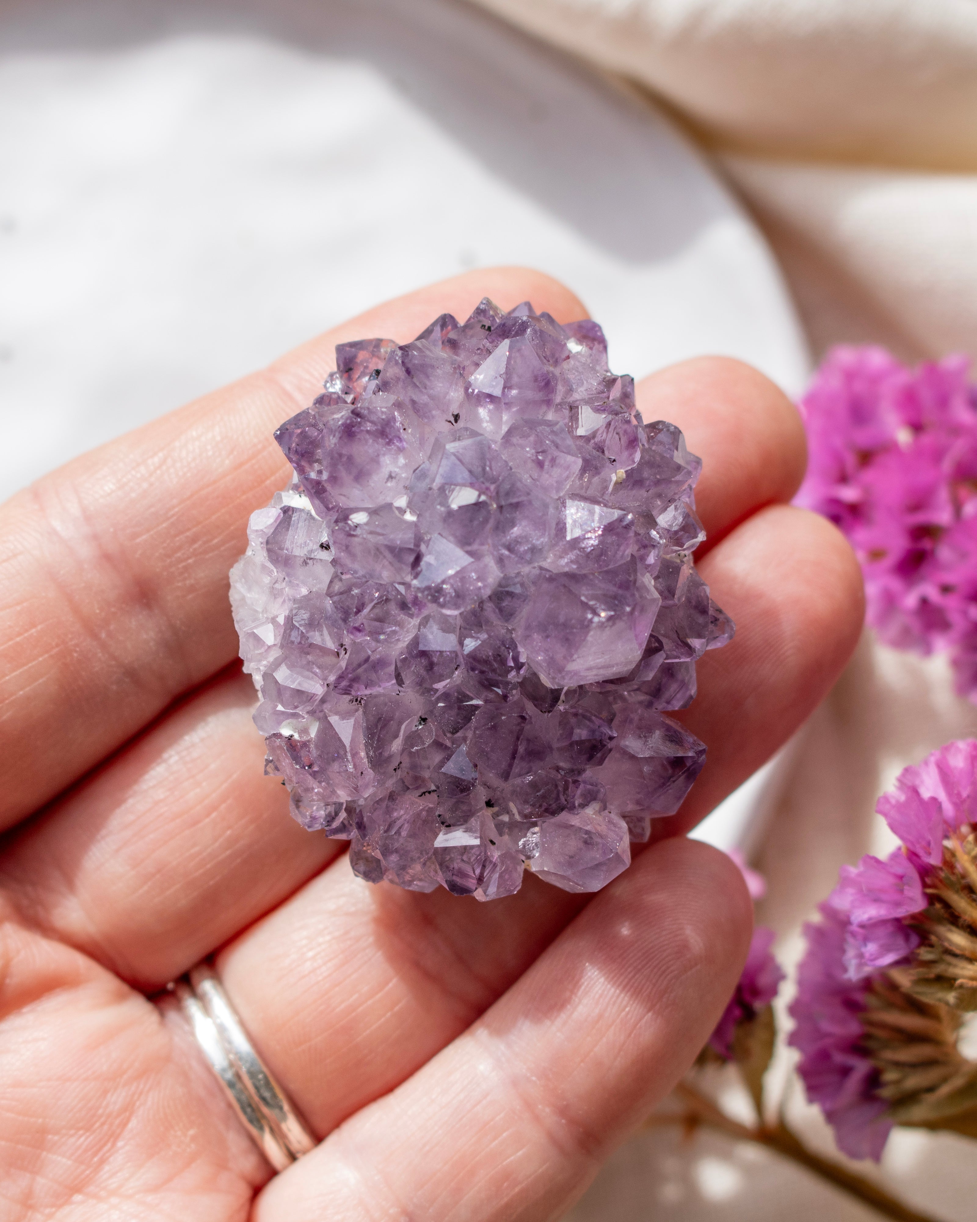 Hand holding a purple crystal with blurred pink flowers in the background