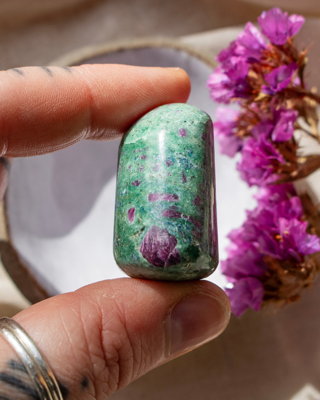 a green and purple stone being held between fingers with purple flowers in the background