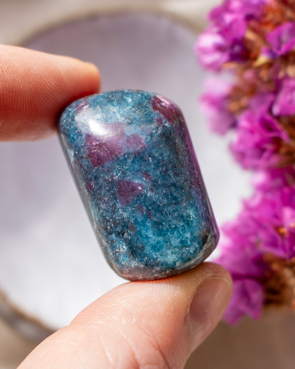 a dark blue and purple stone being held between fingers with a ceramic dish and purple flowers in the background