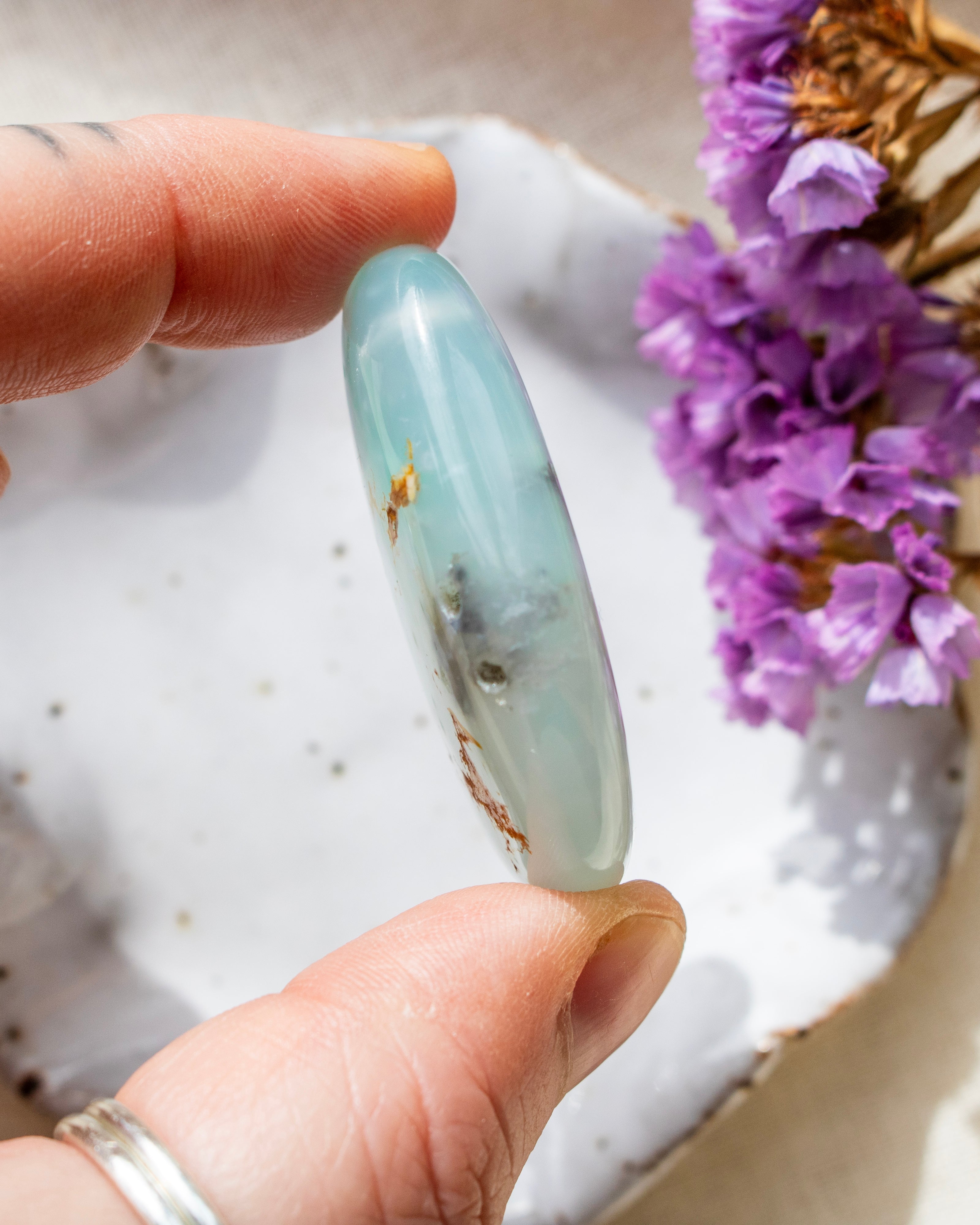 a soft blue stone with rounded edges being held with fingers and a white ceramic dish with purple flowers in the background