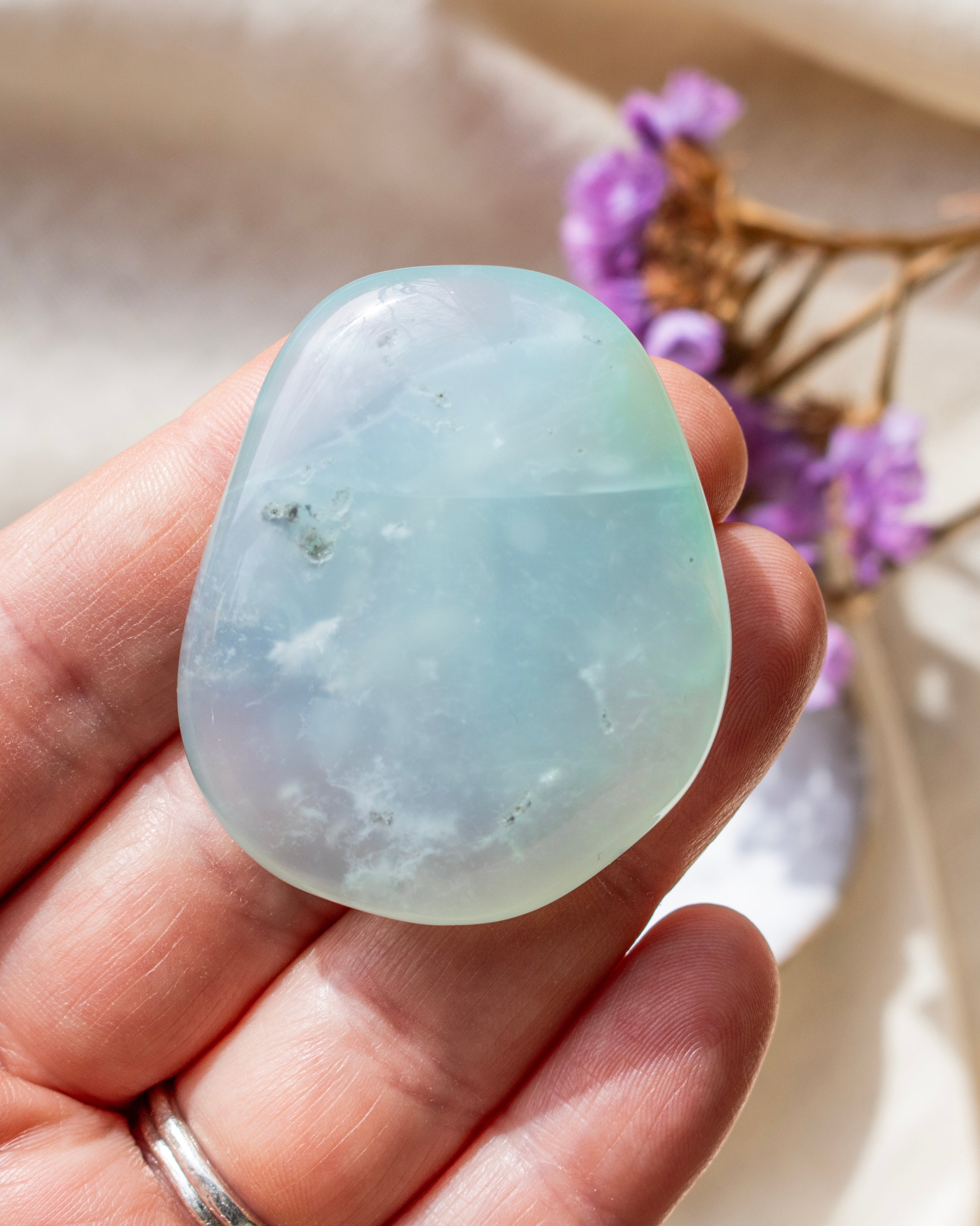 a soft blue stone with rounded edges being held with fingers and a white ceramic dish with purple flowers in the background