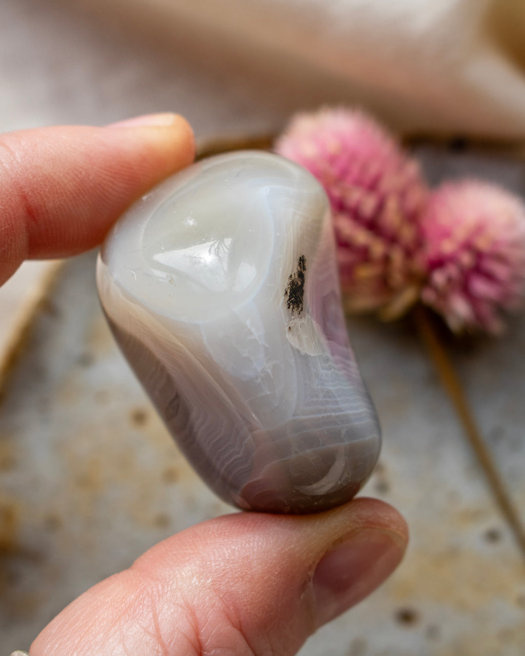 a banded agate pebble being held with fingers and a brown ceramic dish with a pink flower in the background