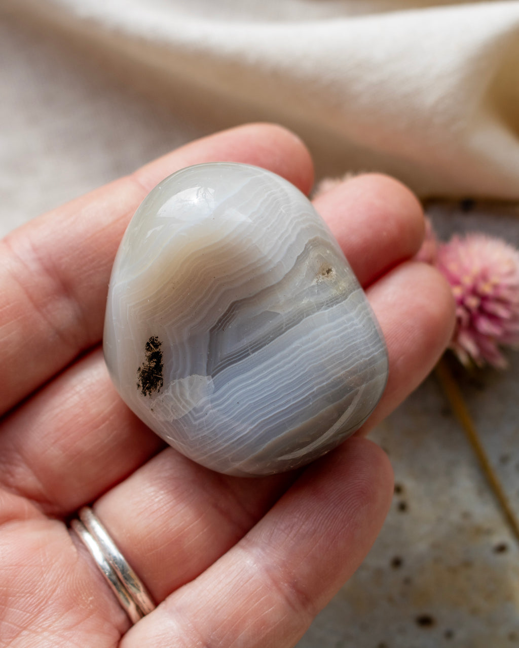 a banded agate pebble being held with fingers and a brown ceramic dish with a pink flower in the background
