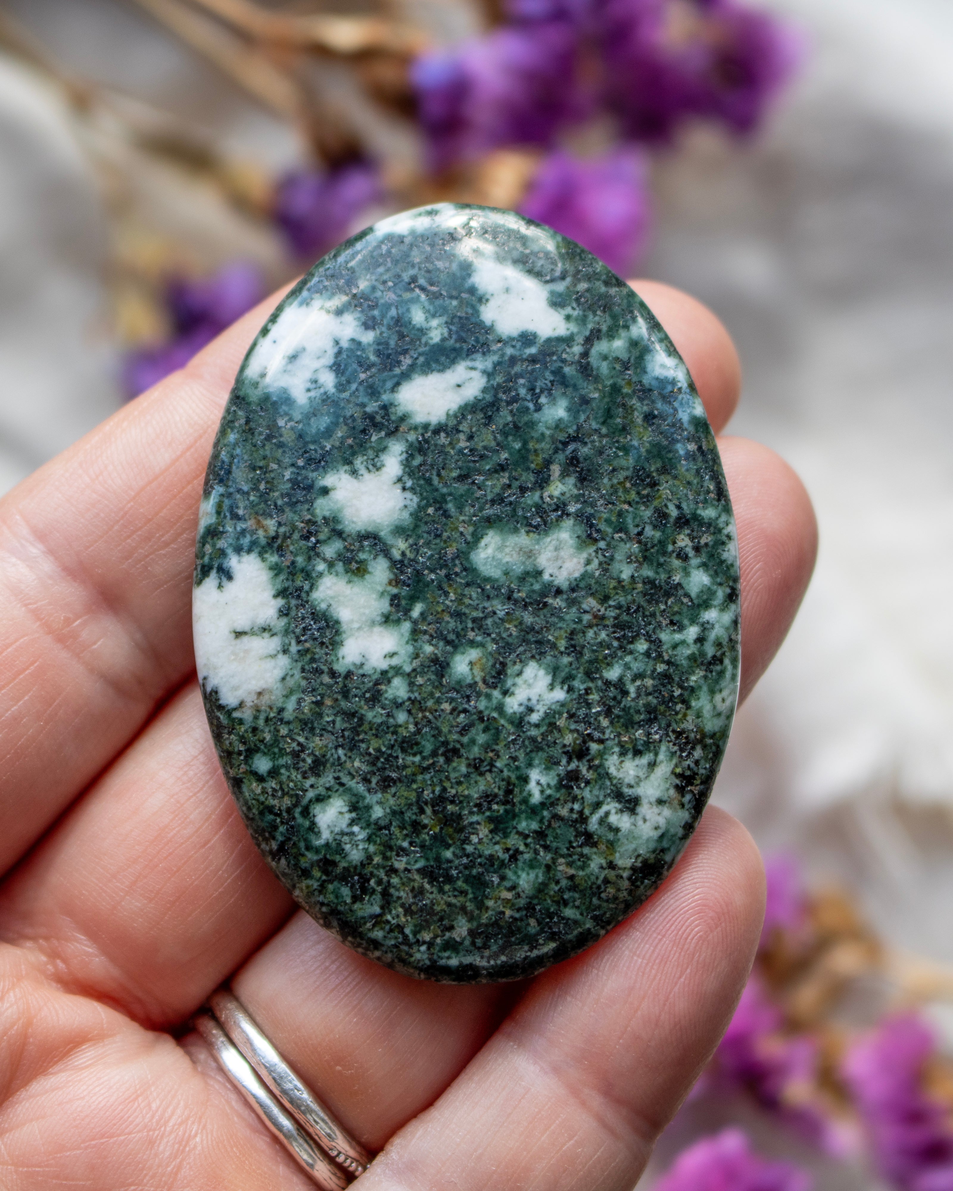Oval-shaped green stone held in a hand with a blurred purple flower background
