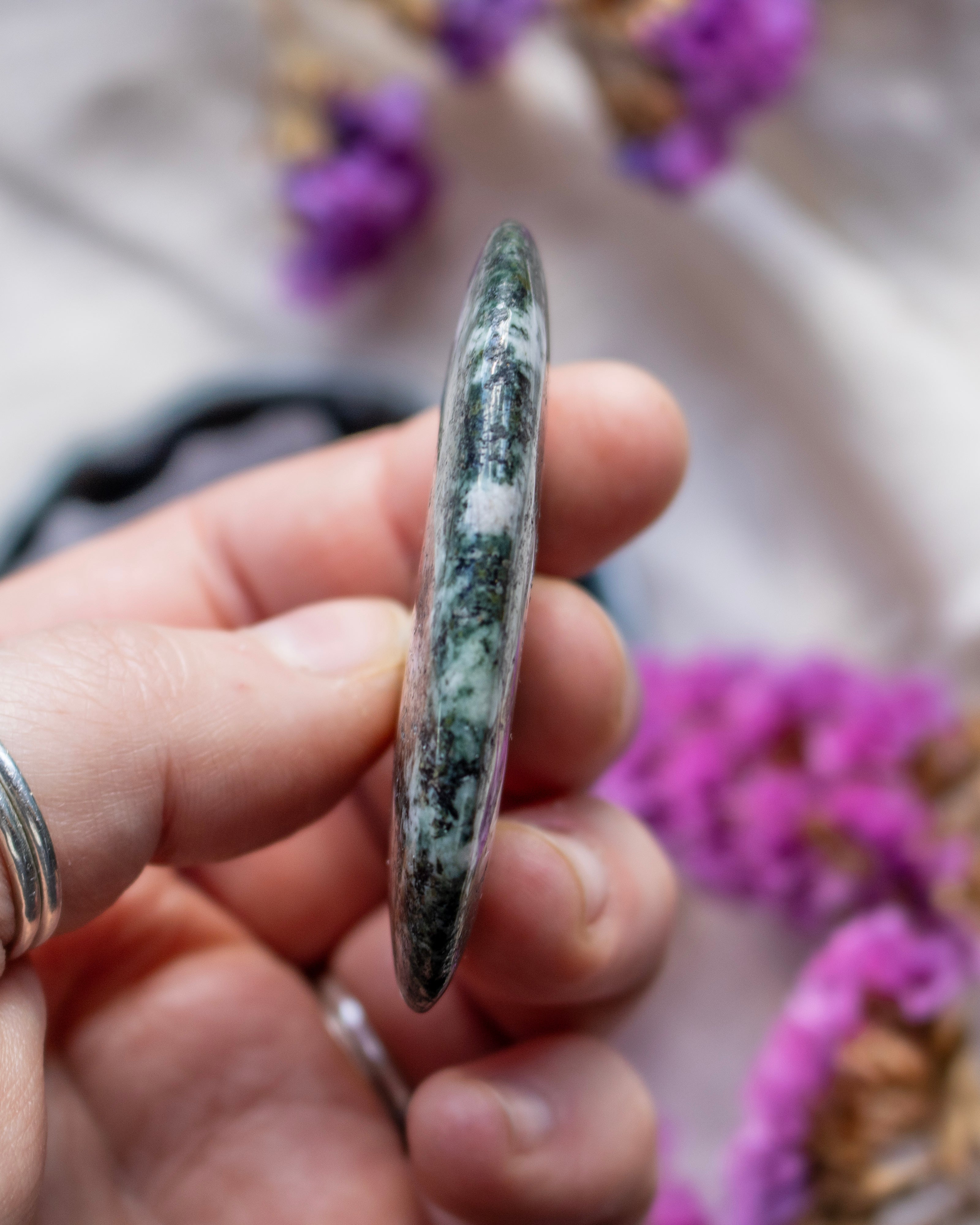 Hand holding a green and white stone with blurred purple flowers in the background