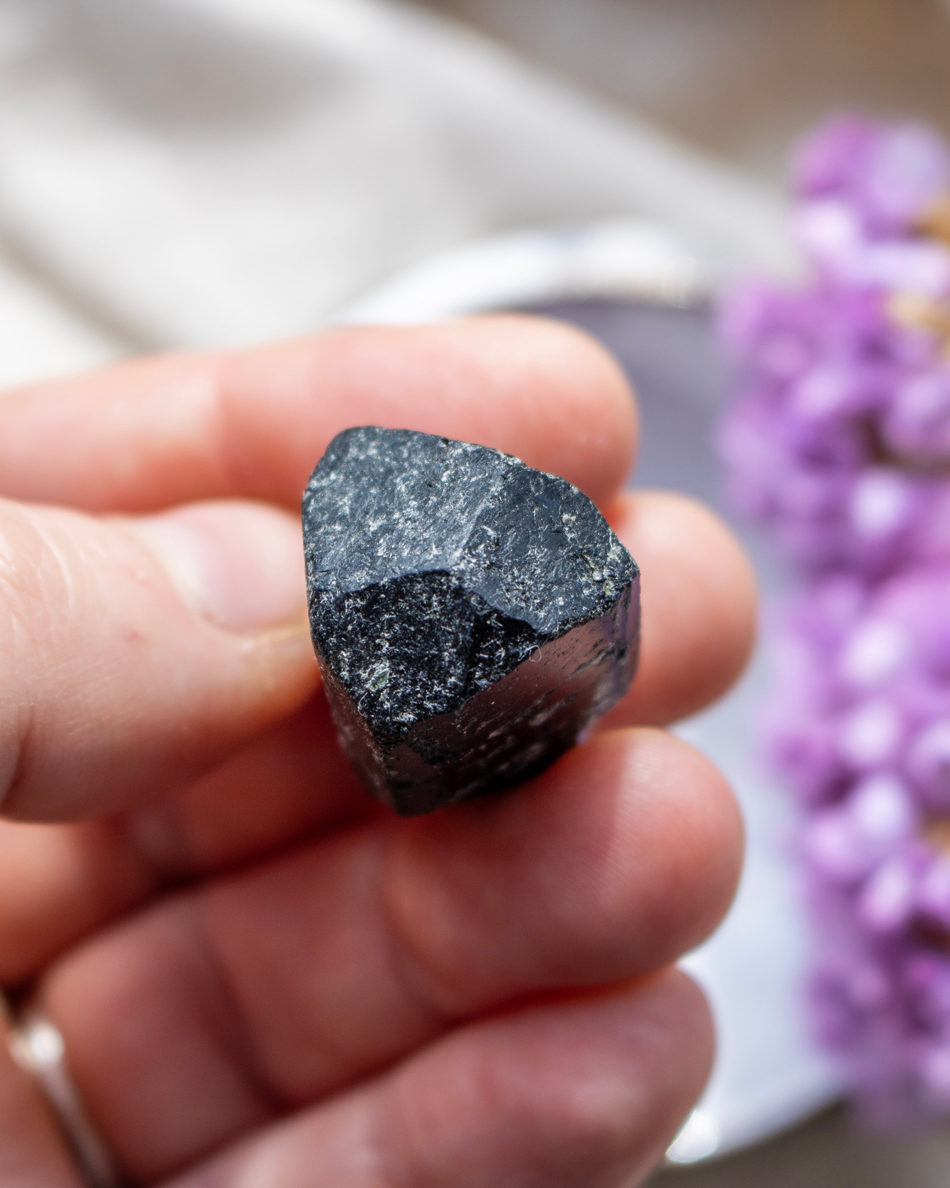 Hand holding a dark crystal with blurred purple flowers in the background