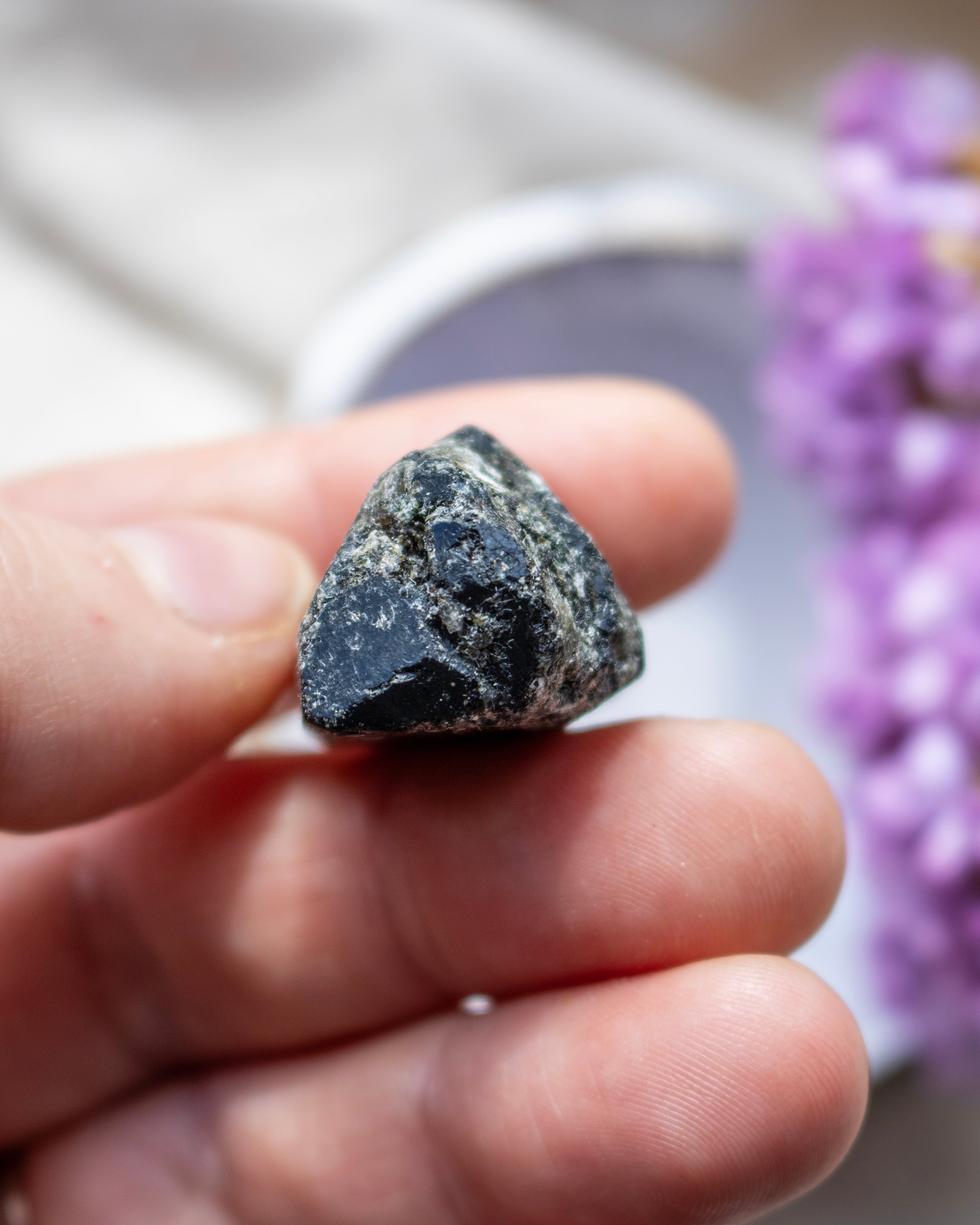 Hand holding a dark stone with blurred purple flowers in the background