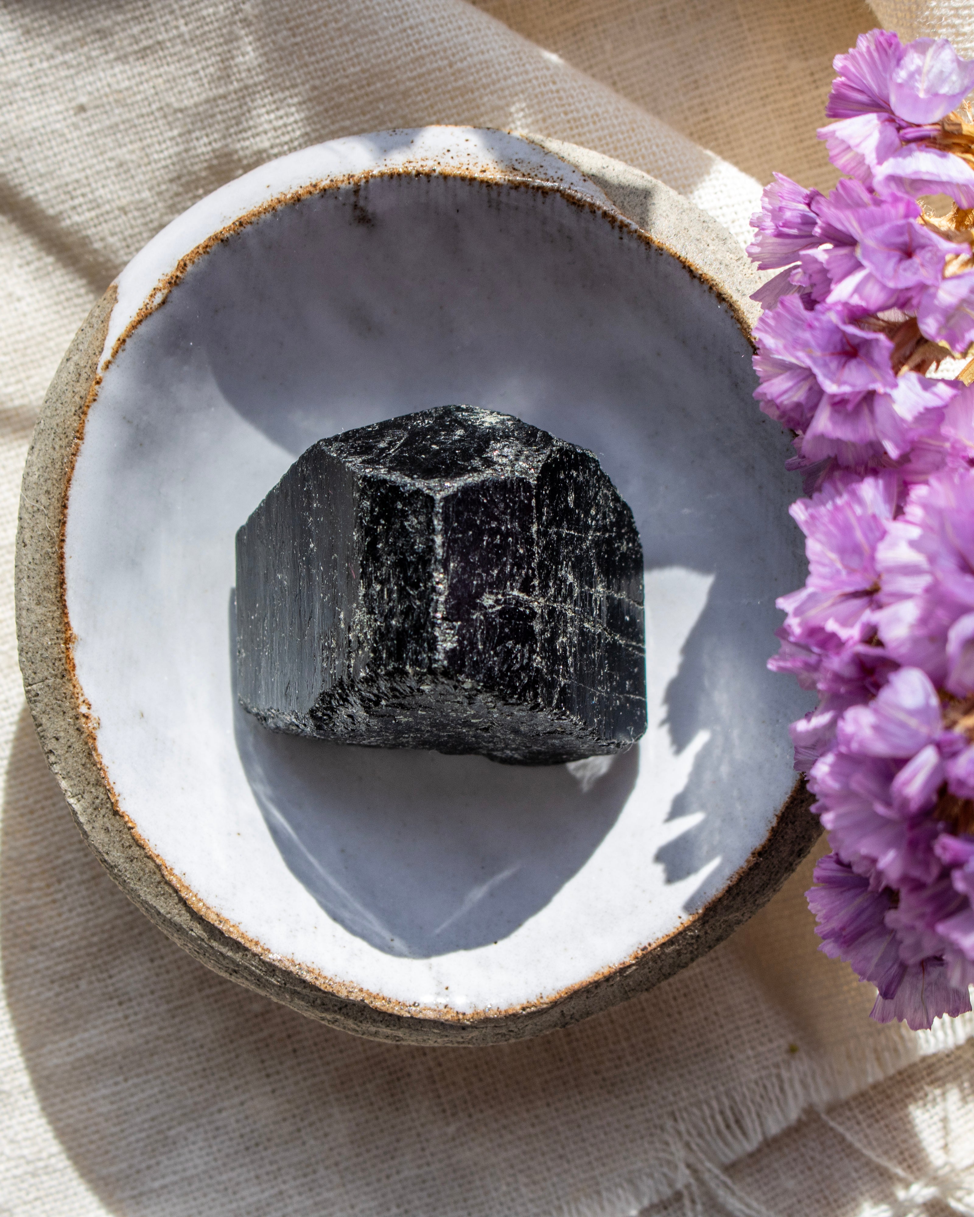 Black stone in a white bowl with purple flowers on a beige fabric background