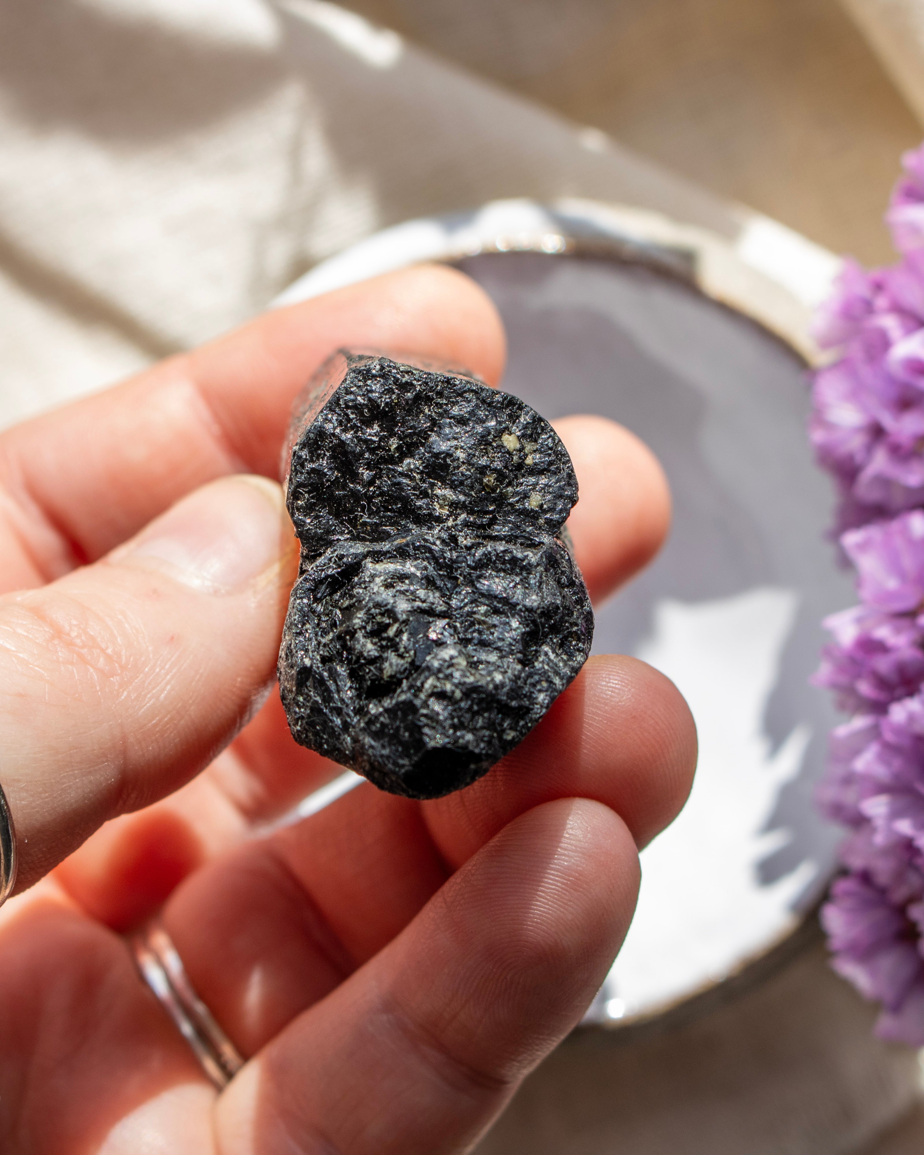 Person holding a dark gray crystal with a blurred background of purple flowers and a neutral surface.