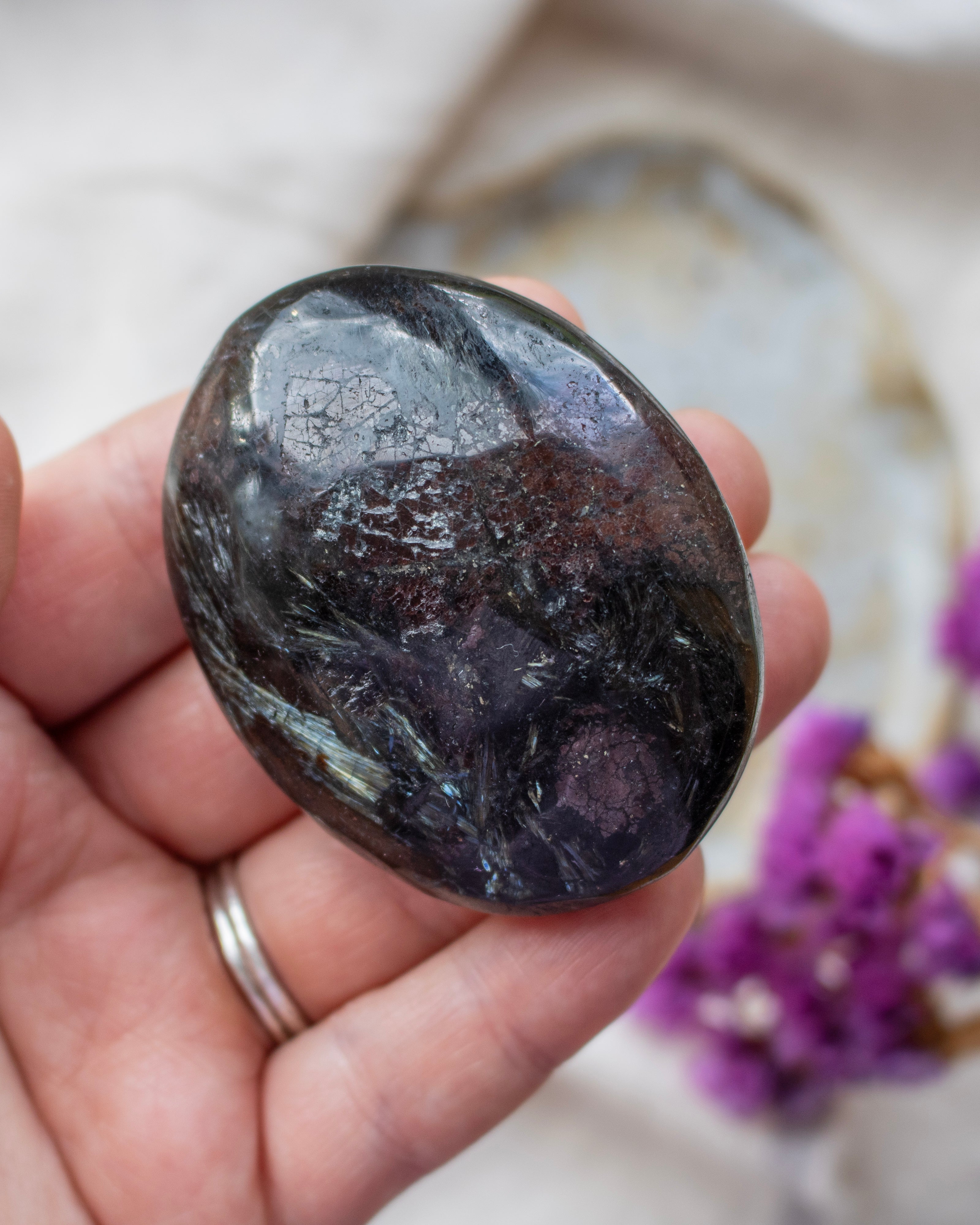 Oval-shaped stone held in a hand with a blurred background featuring purple flowers.