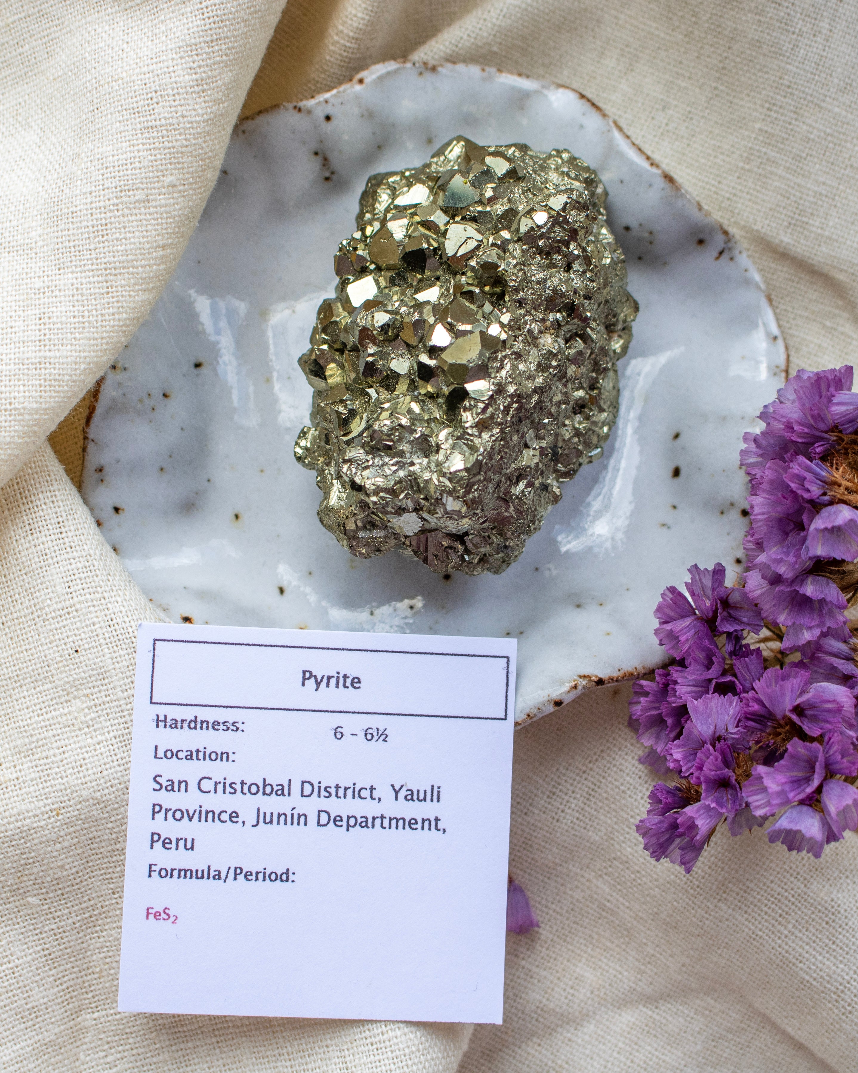 Pyrite crystal on a white stone with a card detailing its properties, placed on a beige fabric background with purple flowers.
