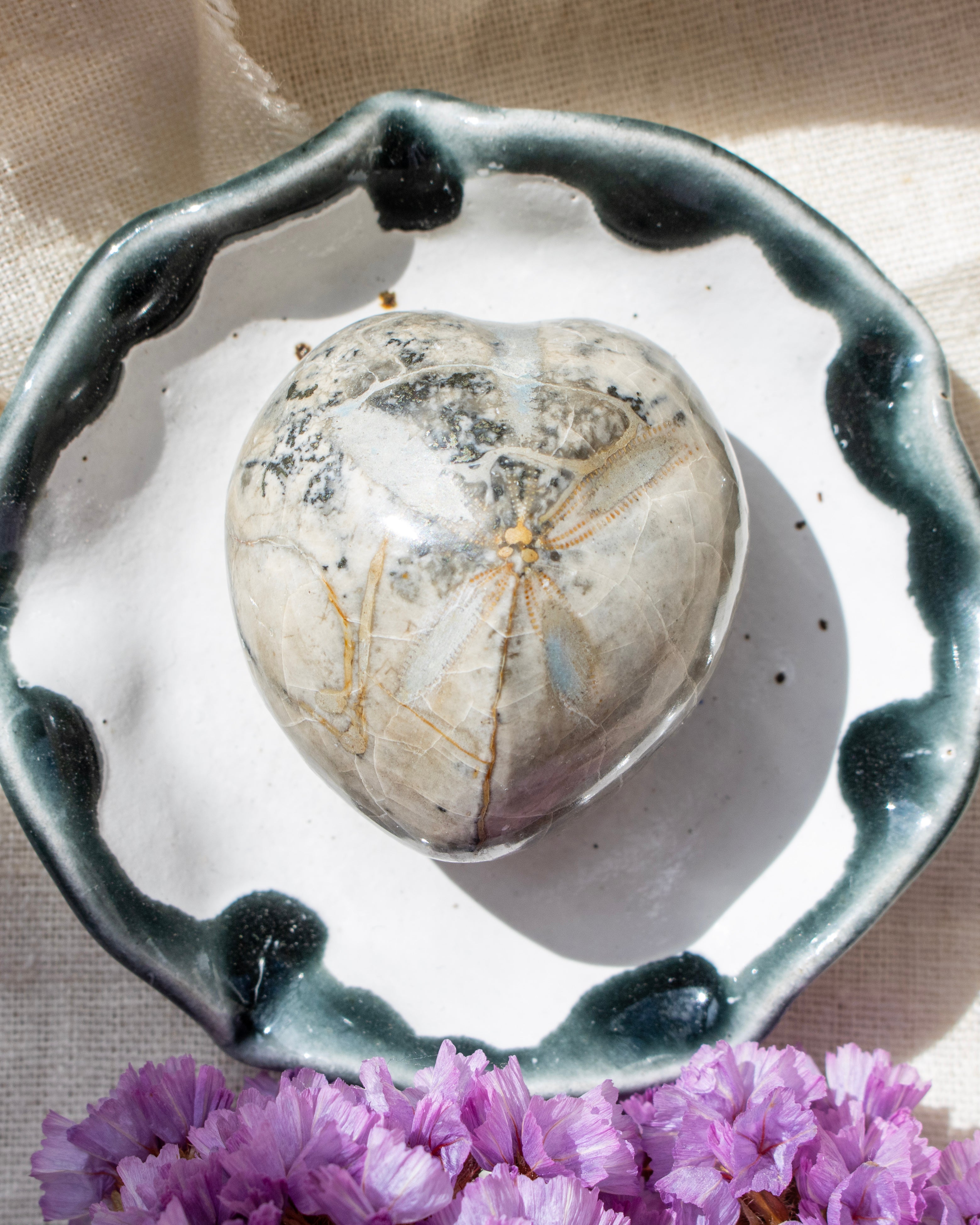 Heart-shaped stone on a white dish with a decorative black rim, surrounded by purple flowers.