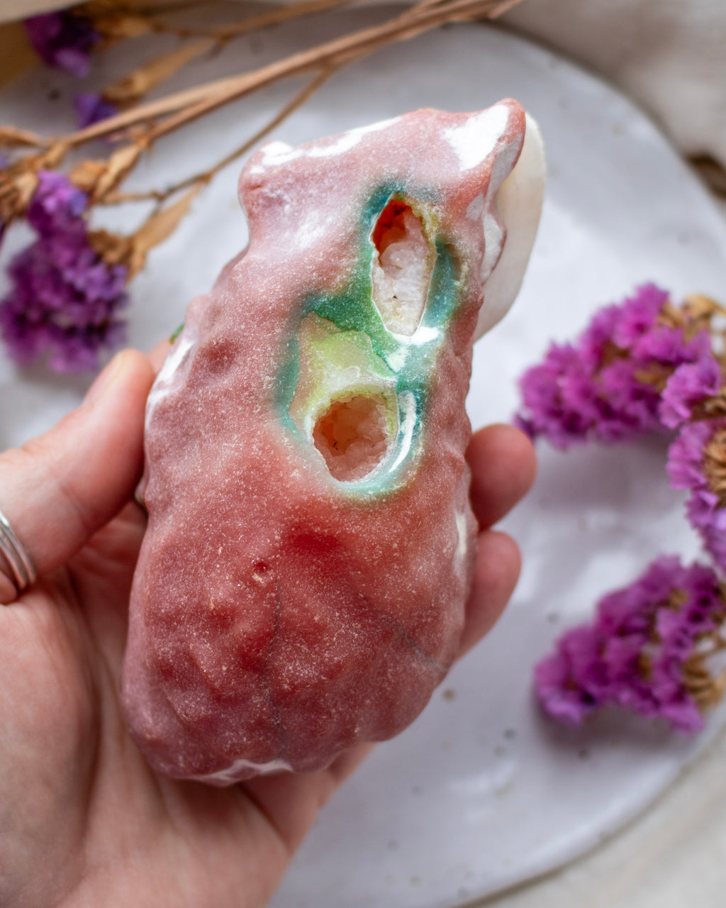 Hand holding a pinkish-brown rock-like object with a hole, on a surface with purple flowers.