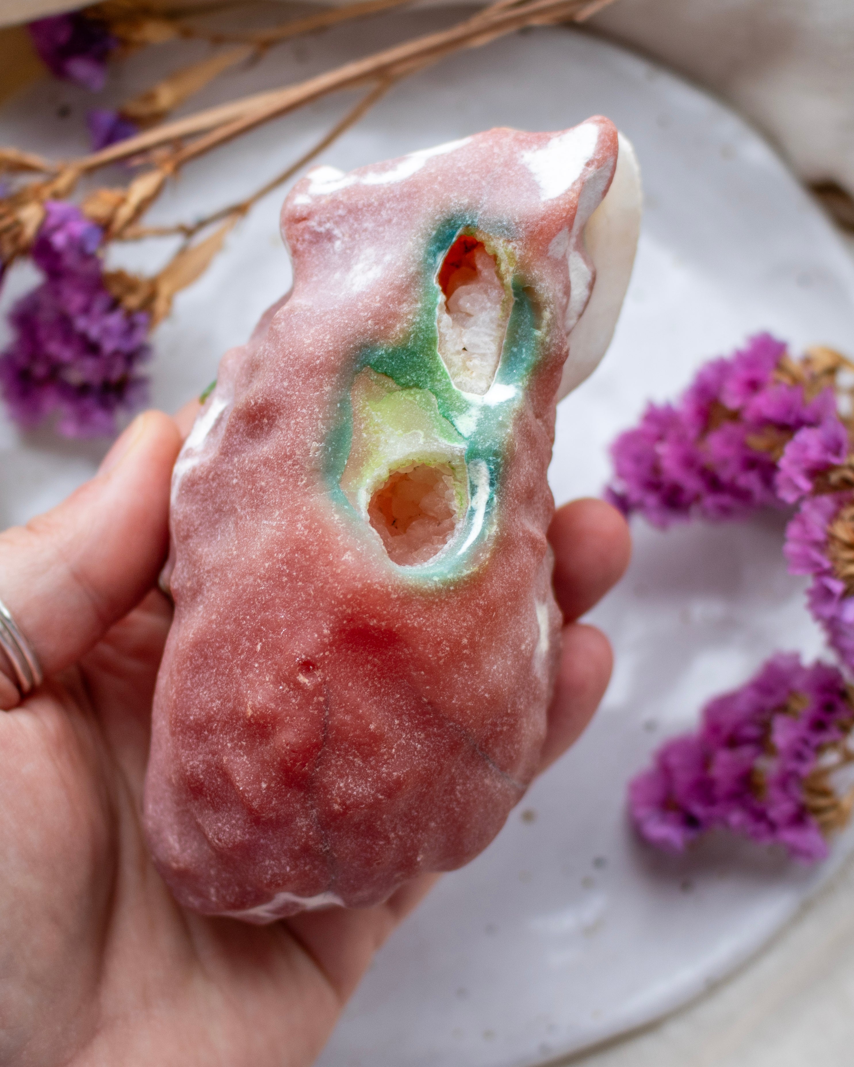 Hand holding a pinkish-brown rock-like object with a hole, on a surface with purple flowers.