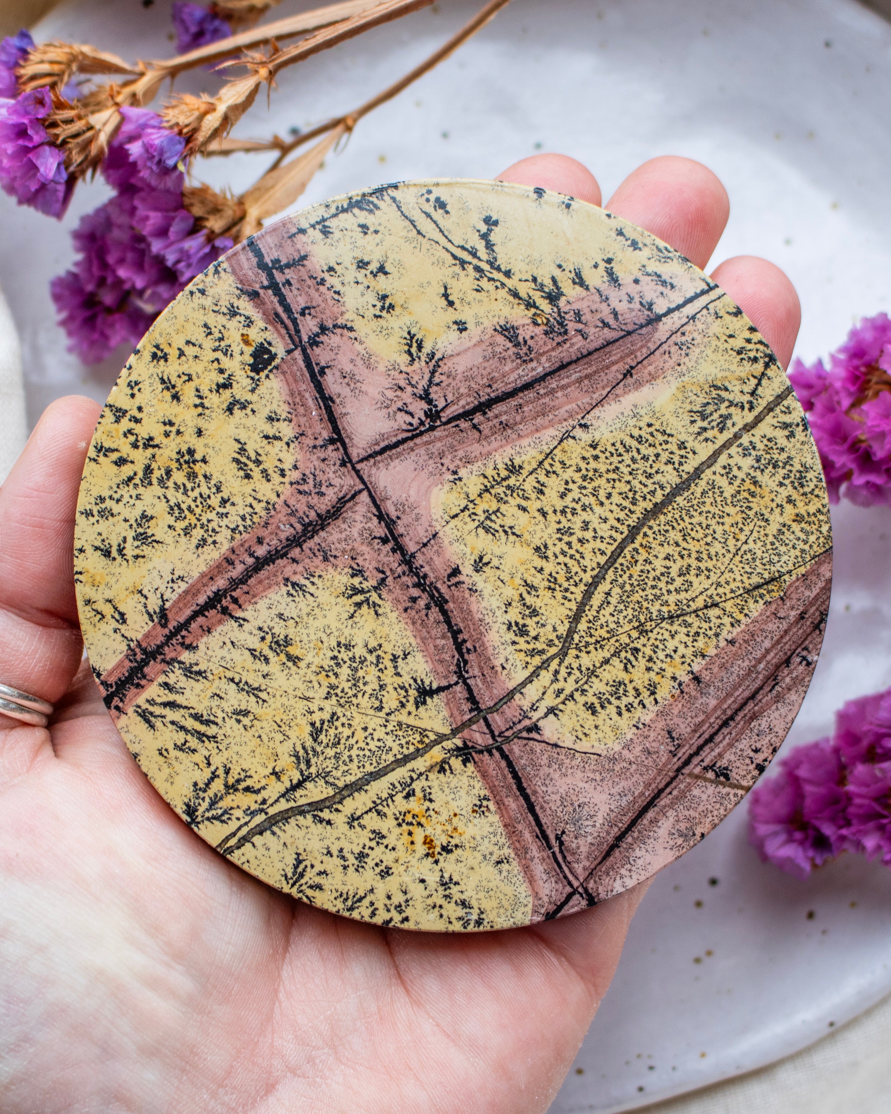 Round stone with leaf pattern held in a hand, surrounded by purple flowers on a light background
