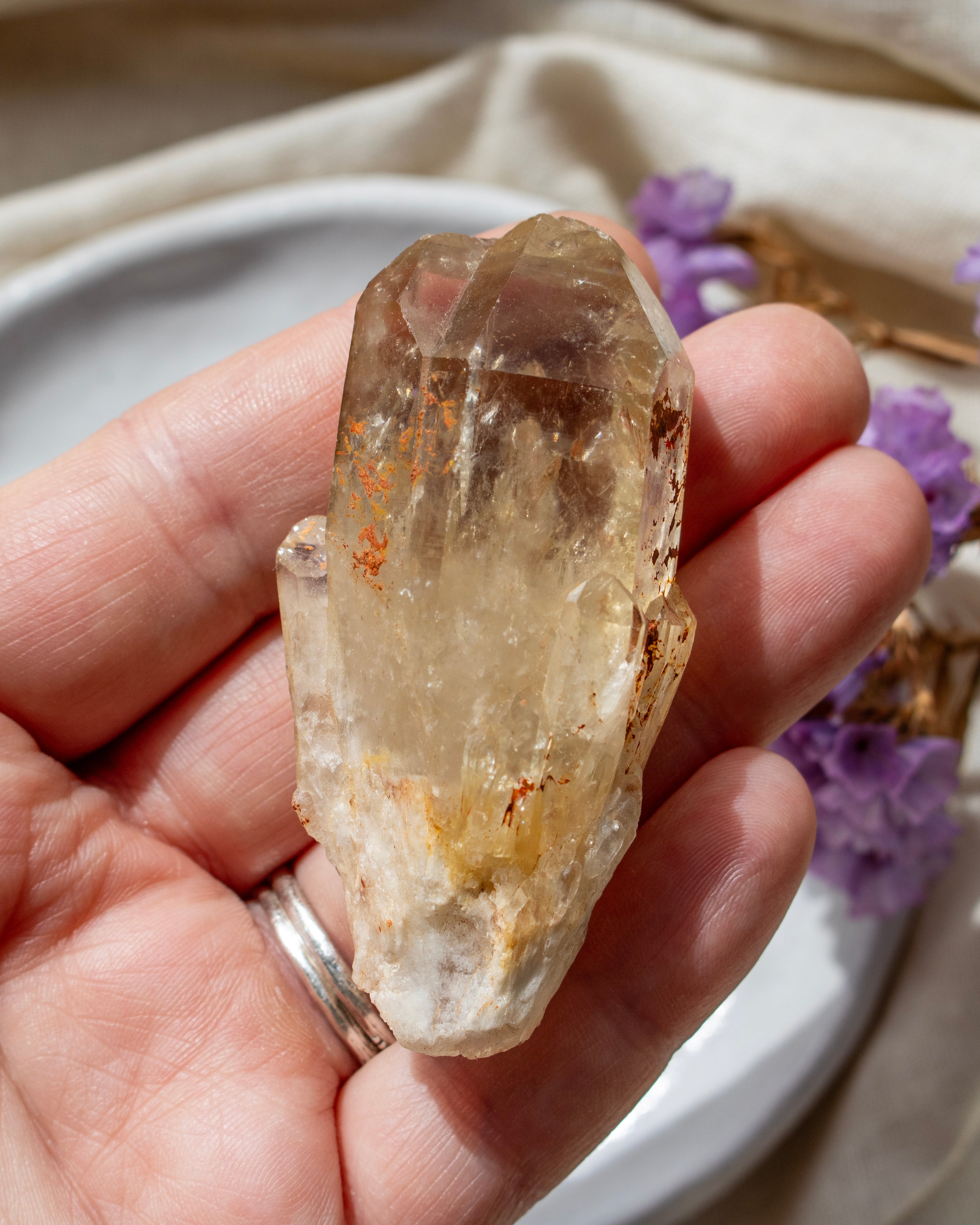 Hand holding a clear crystal with a blurred background of purple flowers and a beige fabric.