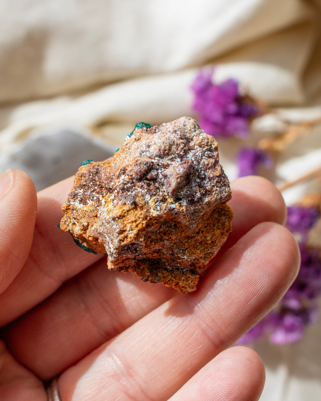 Hand holding a rough mineral specimen with a blurred background of purple flowers and beige fabric.