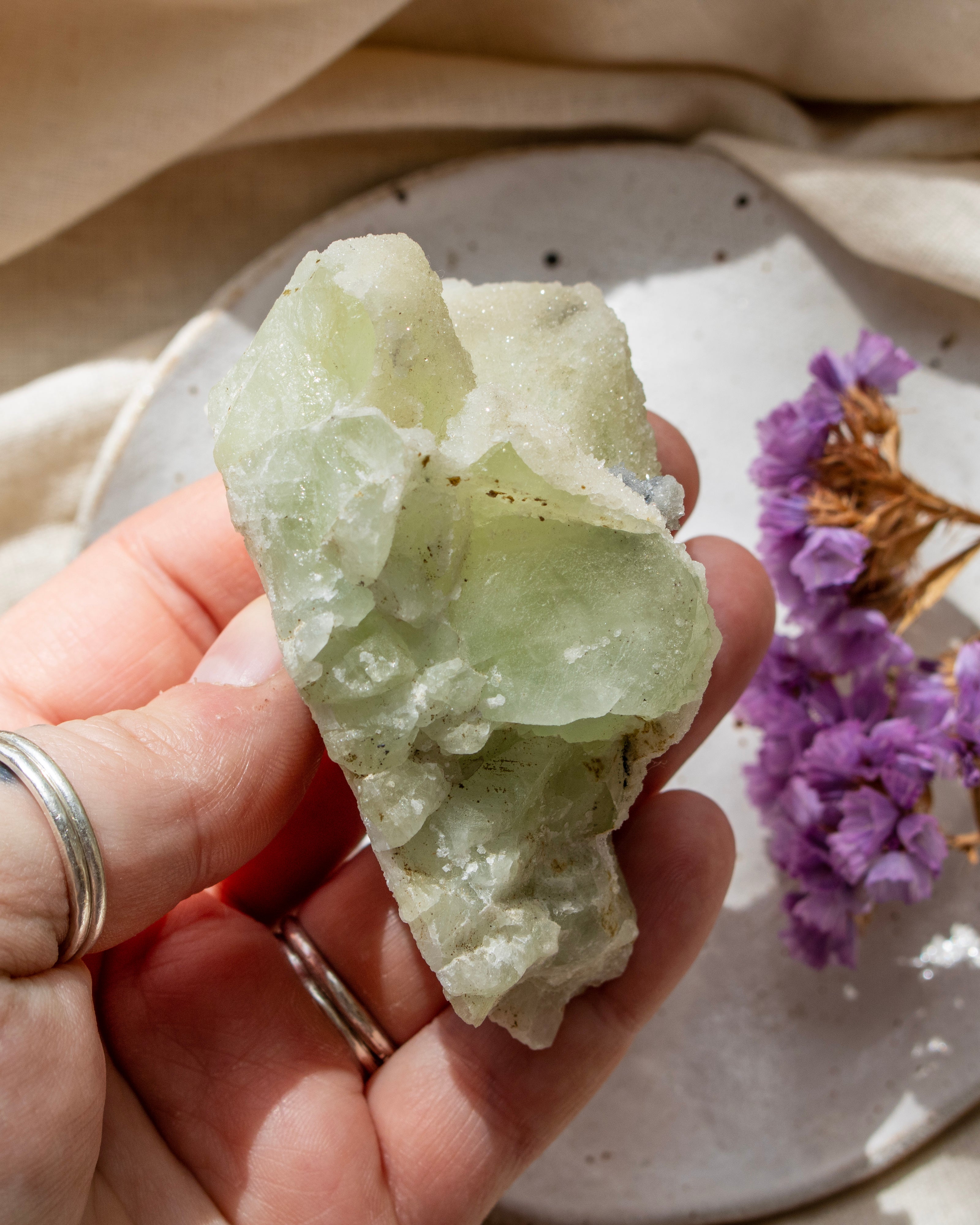 Hand holding a green crystal with a blurred background of flowers and fabric