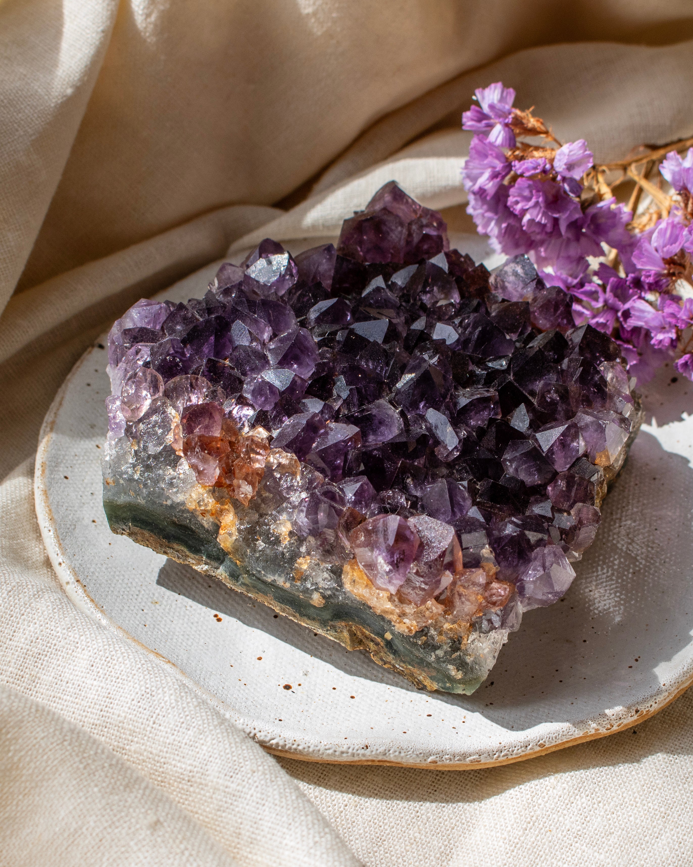 Amethyst crystal cluster on a white plate with purple flowers, beige fabric background