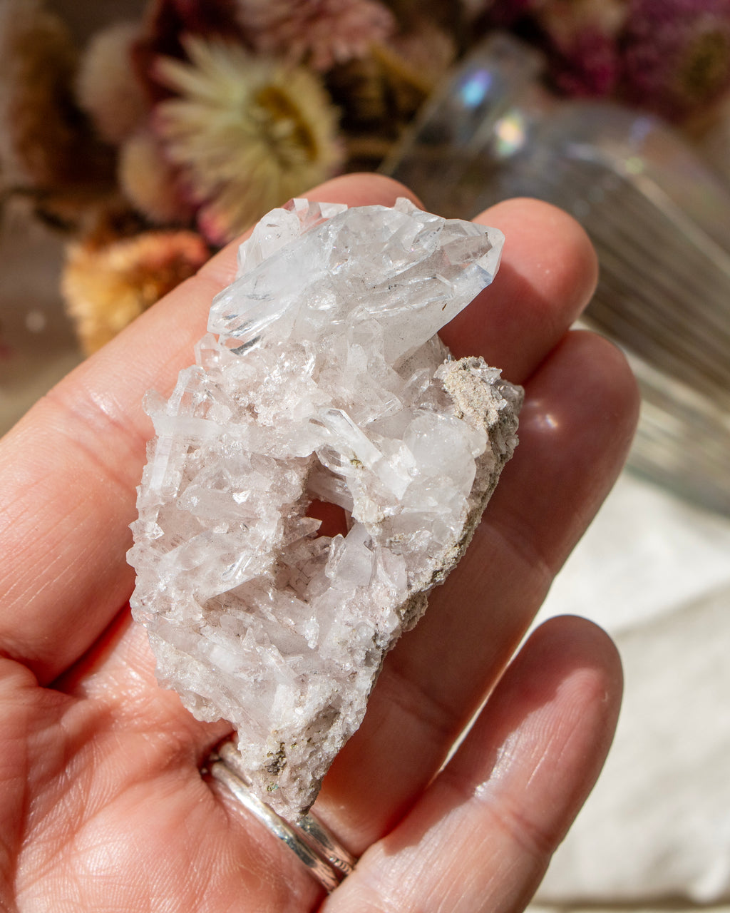 Hand holding a small crystal with a blurred background of flowers and a glass container.