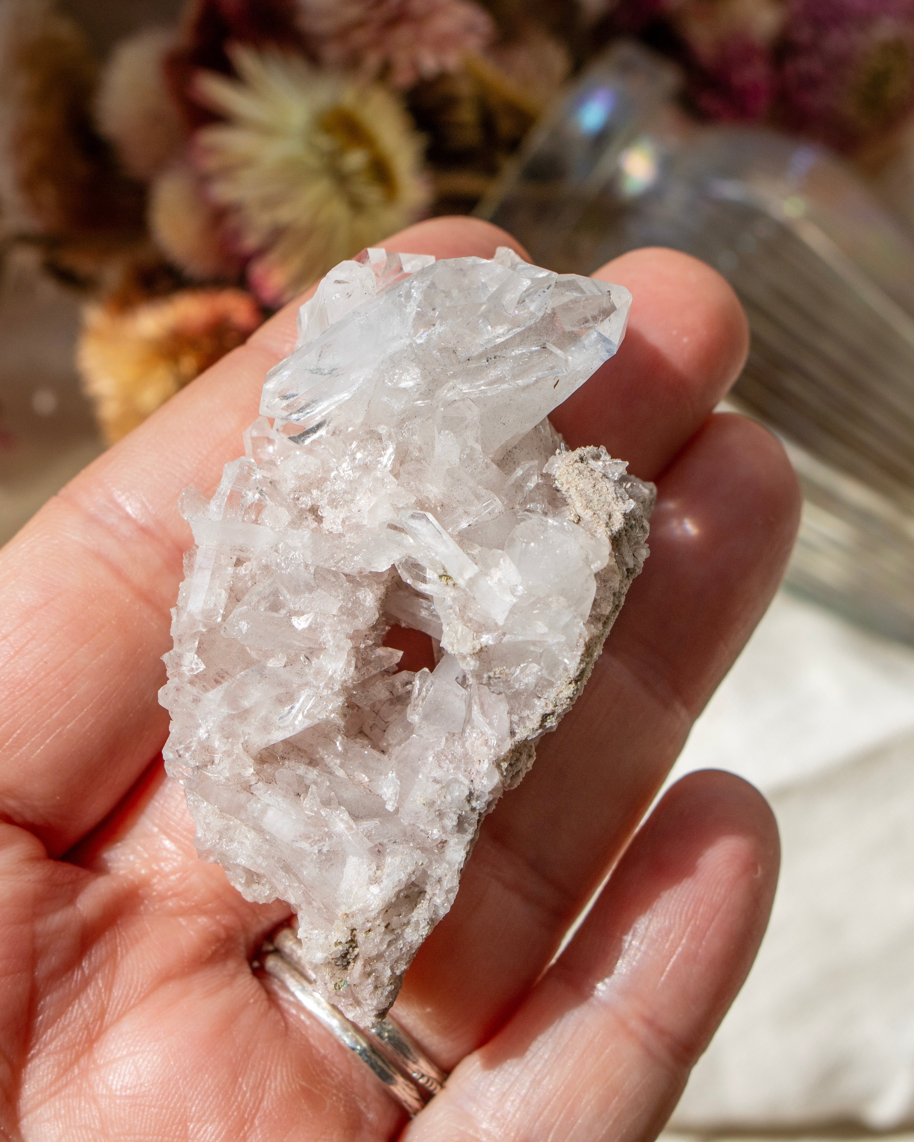 Hand holding a small crystal with a blurred background of flowers and a glass container.