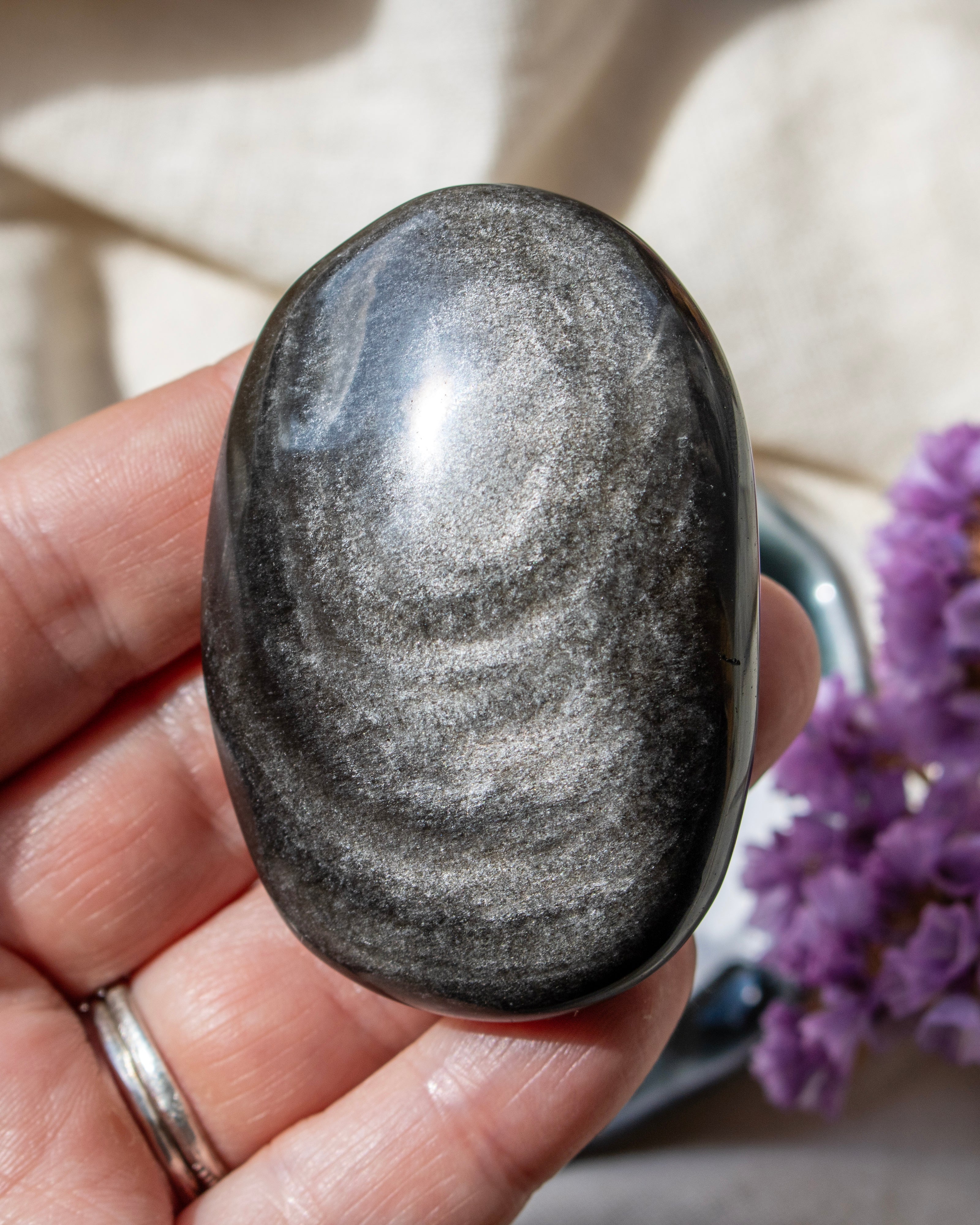 Hand holding a gray stone with a blurred background of purple flowers and a beige surface